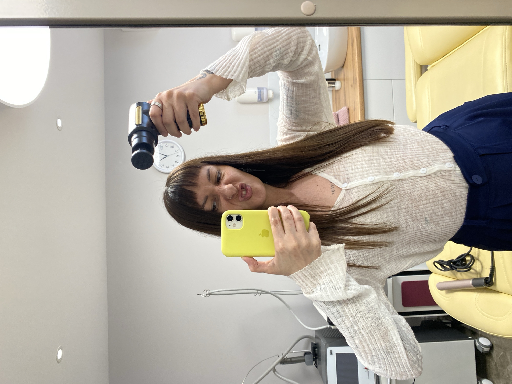 A woman is taking a selfie in a mirror in what appears to be a medical or cosmetic setting. She is holding a yellow iPhone in one hand and a black device, possibly a massage gun, in the other. The woman has brown hair with bangs, and she is wearing a light-colored, button-down shirt. The environment includes a yellow chair, medical equipment, a sink, a clock on the wall, and lighting fixtures. The location is unspecified, and there is no evidence to indicate that the image was taken in Votkinsk, Russia.
