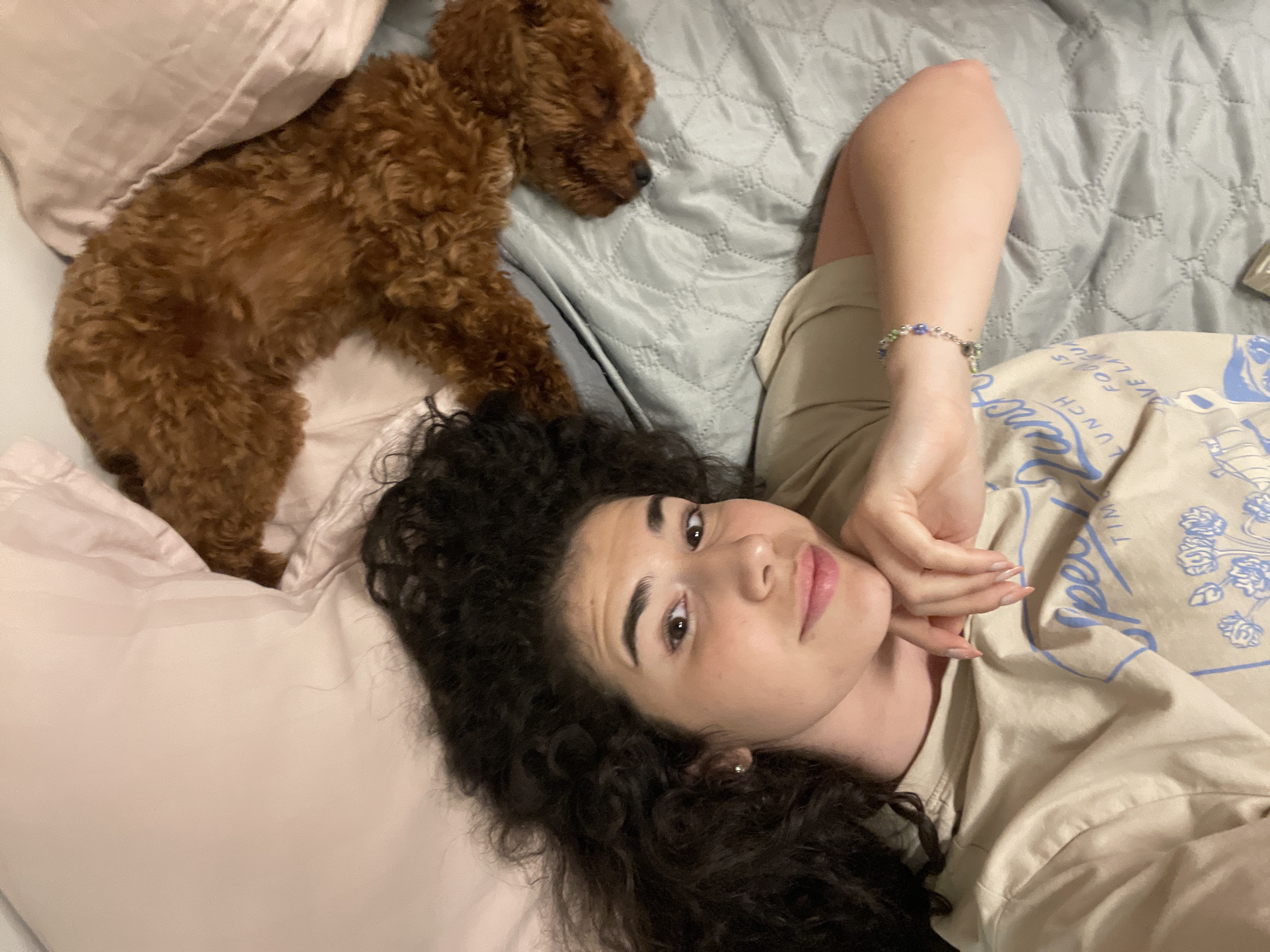 The image depicts a young woman with dark, curly hair lying on a bed. She is wearing a light-colored t-shirt with a blue graphic. Her right arm is bent, and her hand is resting near her face. She is wearing a bracelet with green and purple beads. Next to her lies a small, fluffy, reddish-brown dog, curled up and partially covered by a pink pillow. The bed appears to be covered with a light grey, textured comforter. The scene is indoors, suggesting a private residence. There is no visible indication of the specific location within the city of Sofia, Bulgaria.