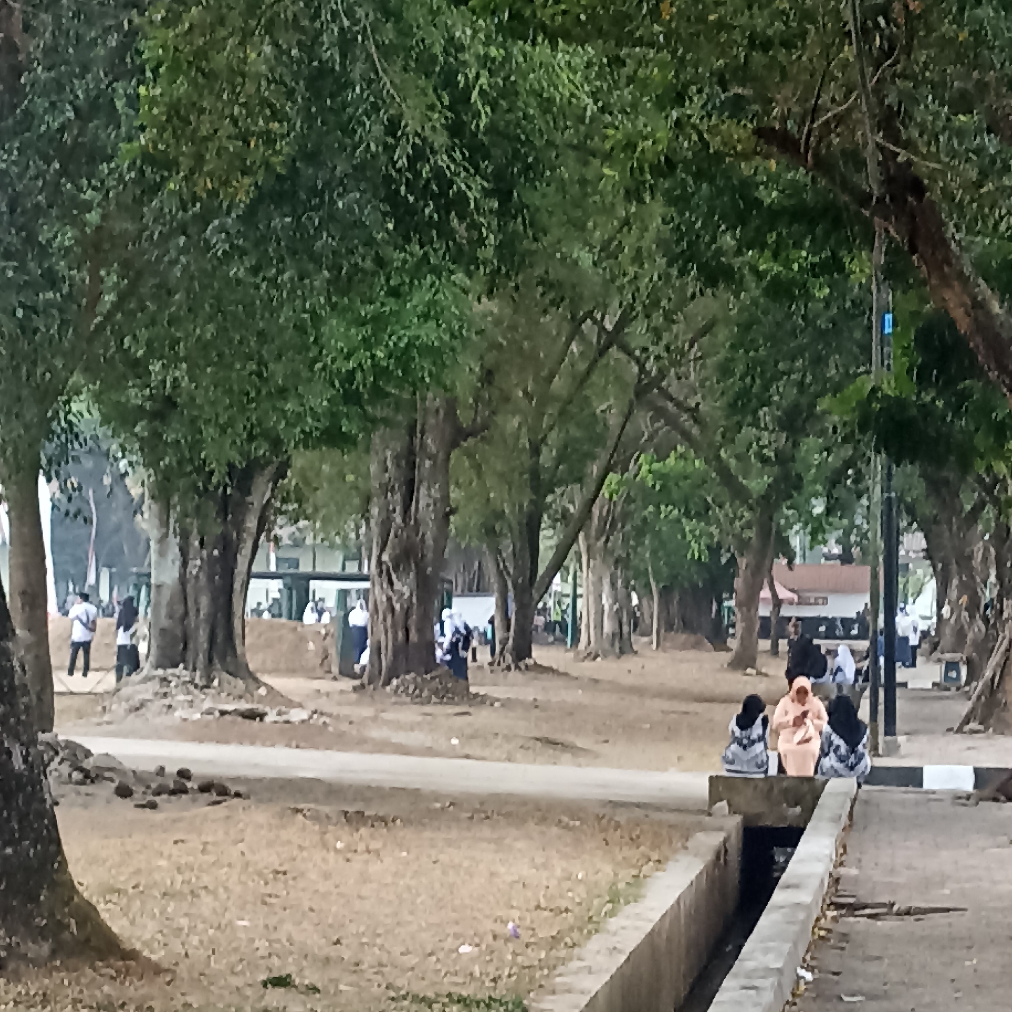 The image depicts a treelined walkway in Magelang, Indonesia, likely within a public park or university campus. Several large trees with dense foliage create a canopy over a dirt and grass area, with a paved path running through it. In the foreground, a concrete channel or drainage ditch runs along the right side of the image. Several people are visible in the scene. On the right, three individuals are seated on a low concrete ledge. One person in the center wears a light orange hijab and light-colored clothing, while the two individuals on either side wear dark hijabs and patterned clothing. Further in the background, more people are scattered throughout the park, some walking and others standing. They appear to be dressed in casual attire and some wear hijabs, consistent with the local culture. The overall atmosphere is one of a relaxed outdoor gathering, possibly during a daytime event or daily activity. The environment suggests a warm climate with the presence of lush greenery.