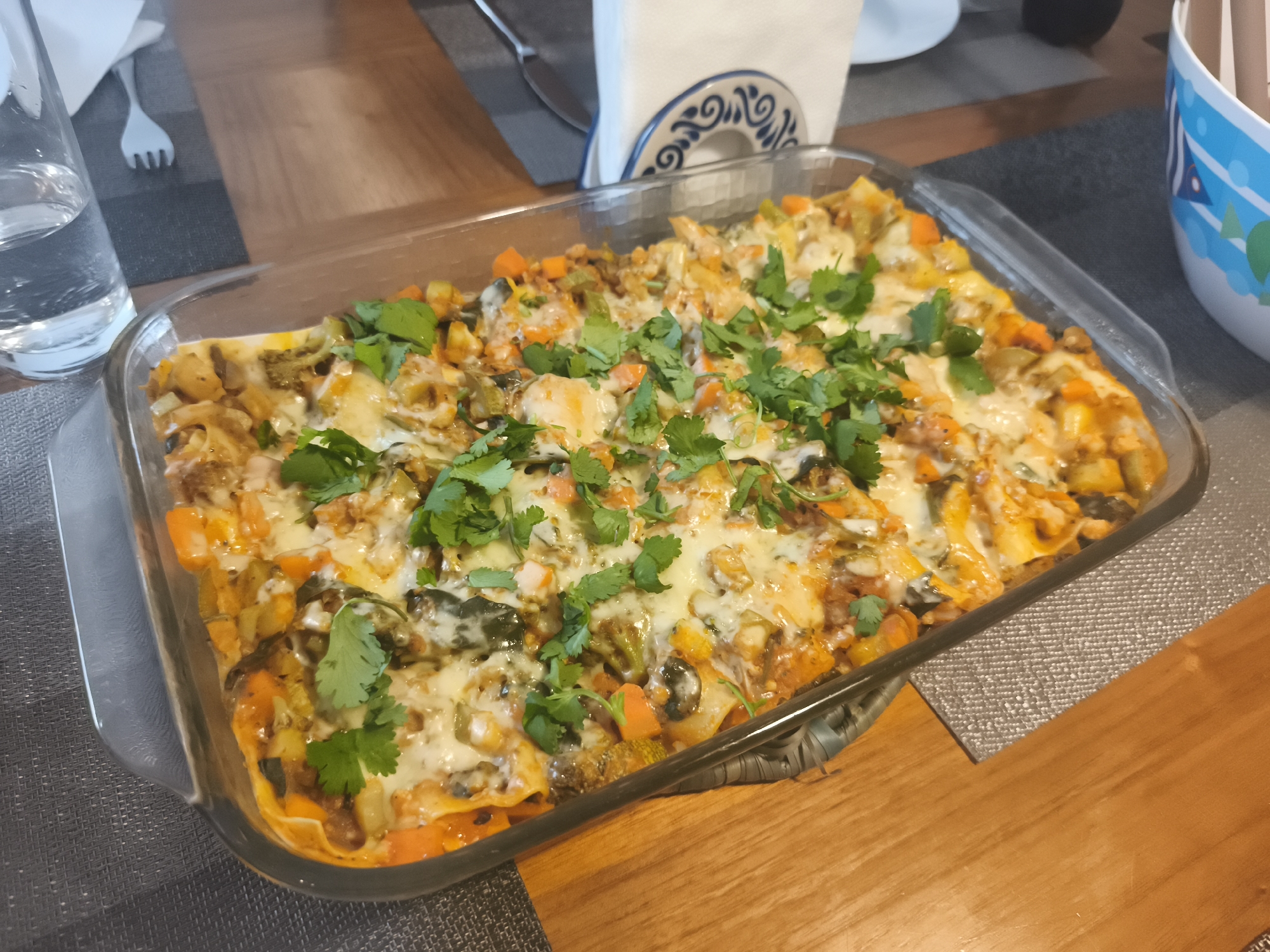 The image shows a glass baking dish filled with a lasagna-like dish topped with melted cheese and fresh cilantro. The dish contains visible layers of pasta, vegetables (including carrots, zucchini, and spinach), and a sauce. It sits on a gray placemat on a wooden table. In the background, there is a glass of water, a fork, a napkin holder with napkins, a white plate, and a bowl with blue and white designs. The photo appears to have been taken indoors, possibly in a dining room. There is no specific information to indicate that this image was taken in Mexico City, Mexico.