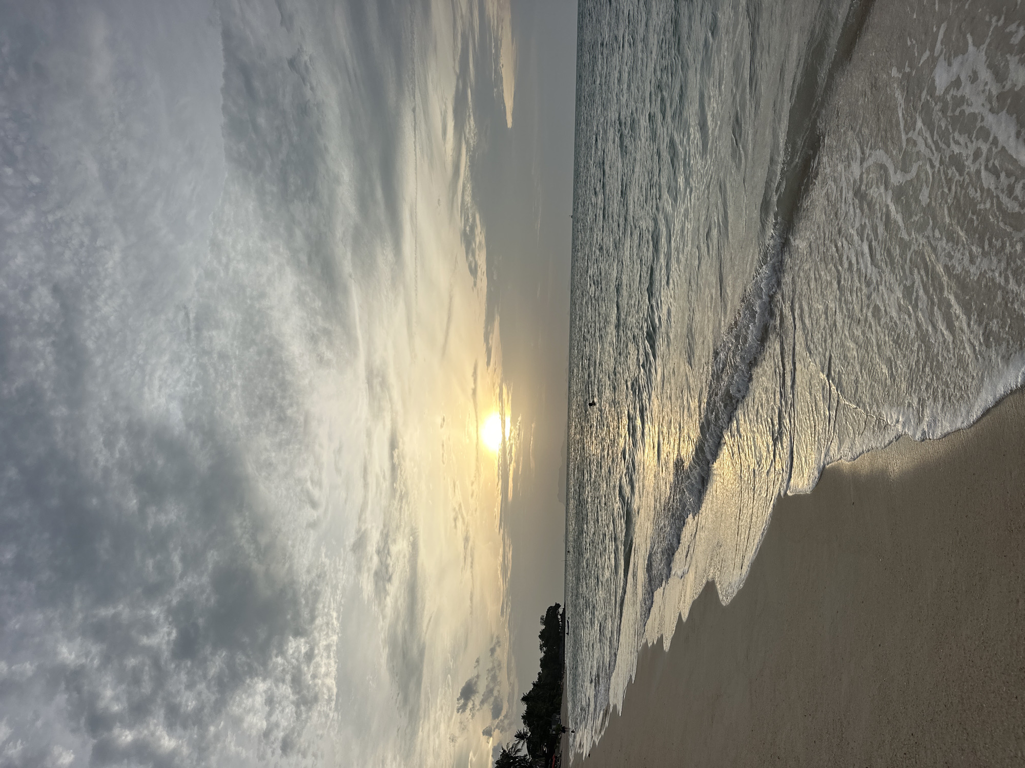 This is a photograph of a beach at sunset on Koh Samui, Thailand. The sun is partially obscured by clouds and is low in the sky, casting a golden glow on the water and sand. The ocean is calm with gentle waves lapping at the shore. The sand is a light beige color, and there are palm trees visible in the distance along the coastline. There are a few people in the water and on the beach, but the scene is generally tranquil. The sky is filled with dramatic, gray clouds.