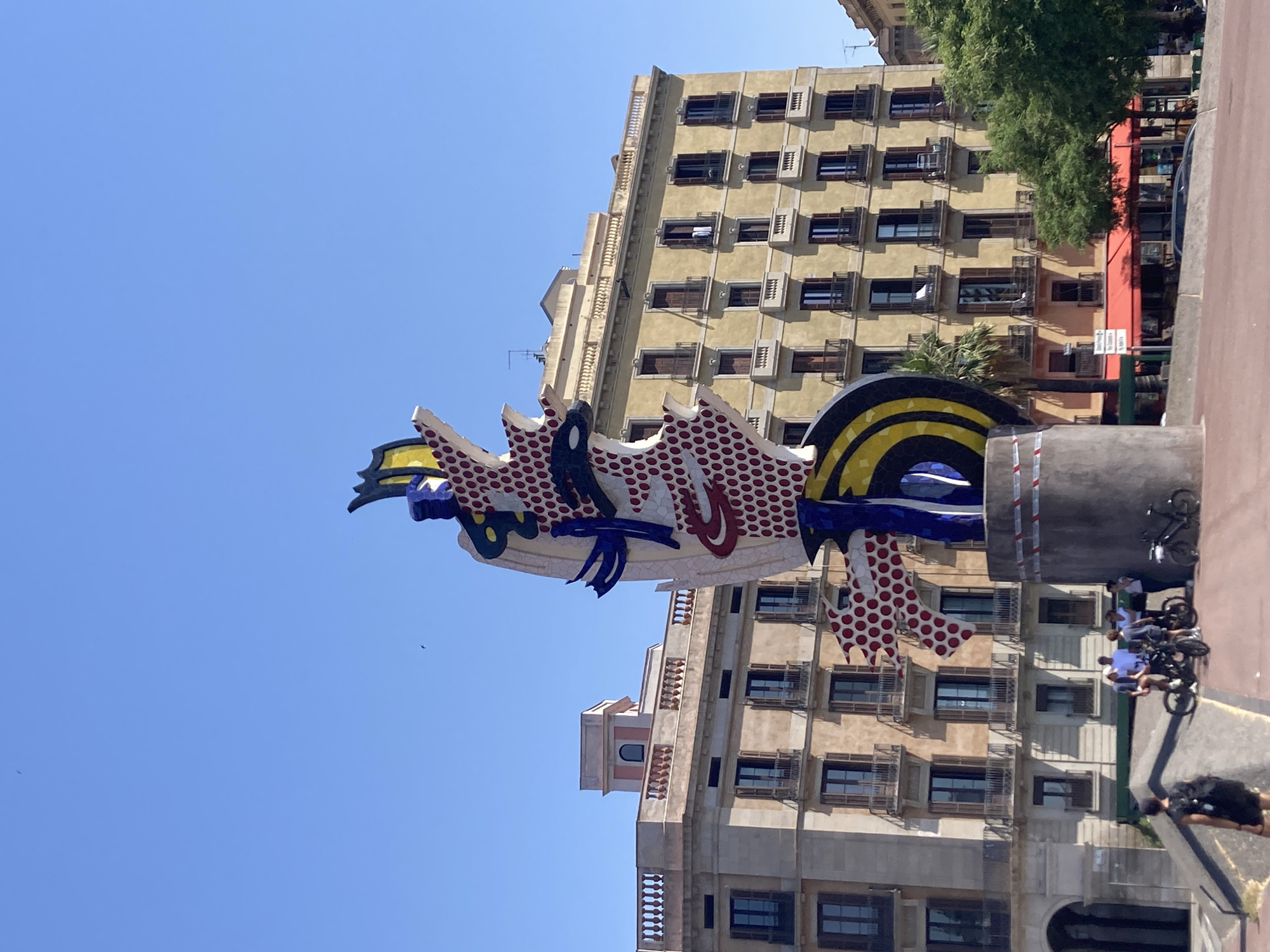 This is a medium-shot photograph taken on a clear, sunny day in Barcelona, Spain. The primary subject is the large, colorful sculpture of a woman's face, known as "La Dona i l'Ocell" (The Woman and the Bird) by Joan Miró, located in Parc de l'Espanya Industrial.
The sculpture is a towering, abstract piece made of mosaic and ceramic tiles. It features a fragmented face with bold colors like red, yellow, blue, and black, and characteristic dotted patterns. The sculpture is situated on a cylindrical concrete base, partially obscured by a red and white striped tape.
Behind the sculpture, two large, multi-story buildings with tan facades and numerous windows are visible. The windows have dark wooden shutters, some of which are open. The buildings have architectural details like balconies with wrought-iron railings and decorative cornices. The sky above is a clear, vibrant blue with a few small birds visible.
In the foreground, several people are gathered near bicycles. Two individuals are standing beside a bicycle, while others are seated on or near bicycles, suggesting a group of cyclists. One person in the lower left corner is walking away from the camera, carrying a backpack.
The ground in the foreground is paved with reddish-brown material. To the right of the sculpture, a large green tree with dense foliage is visible. Below the buildings, awnings of a reddish hue suggest outdoor seating for a cafe or restaurant.