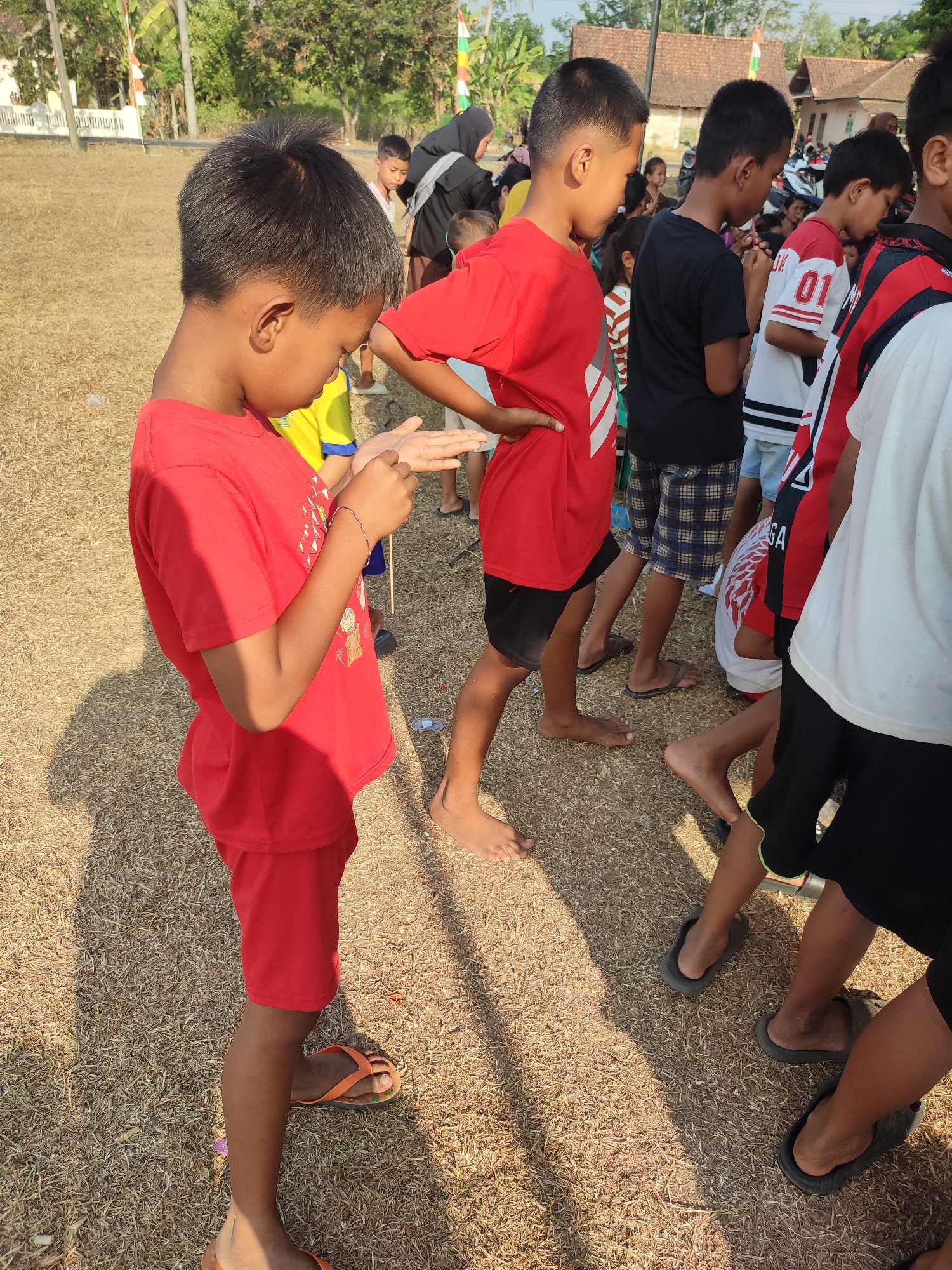 The image depicts an outdoor scene with several children and adults in what appears to be a grassy field. In the foreground, a young boy wearing a red t-shirt and red shorts is holding his hand up, looking down at it. He is wearing orange flip-flops. Behind him, another boy in a red t-shirt and black shorts stands with his hands clasped in front of him. Several other children are visible in the midground, some wearing t-shirts with numbers, and one in a black headscarf. In the background, there are buildings with red-tiled roofs and trees, suggesting a rural or semi-rural setting. The ground is covered with dry grass and straw. The lighting suggests it is daytime, with shadows cast on the ground. Based on the presence of Indonesian flags in the background and the overall environment, the location is likely Nusa Jaya, Indonesia.