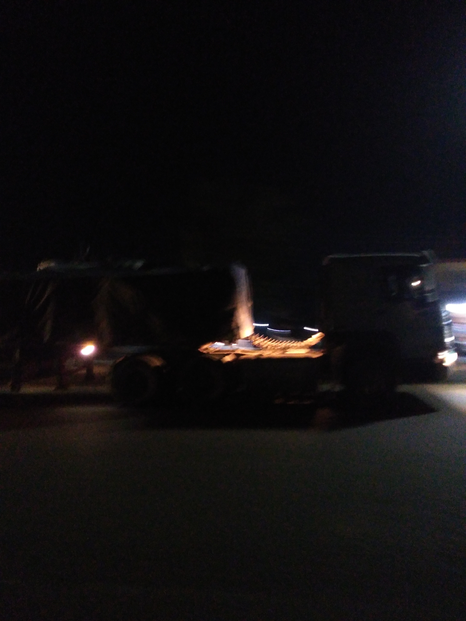 In the city of Jos, Nigeria, a semi-trailer truck is parked at night on an asphalt surface. The truck is dark in color and appears to be covered with a tarp. The image is blurry due to low light conditions and movement. The truck has large wheels and a long trailer. There are lights visible on the truck, including headlights and tail lights, as well as some illuminated elements on the trailer. The background is dark, suggesting it is nighttime. The scene appears to be an outdoor setting, possibly a road or a parking area.