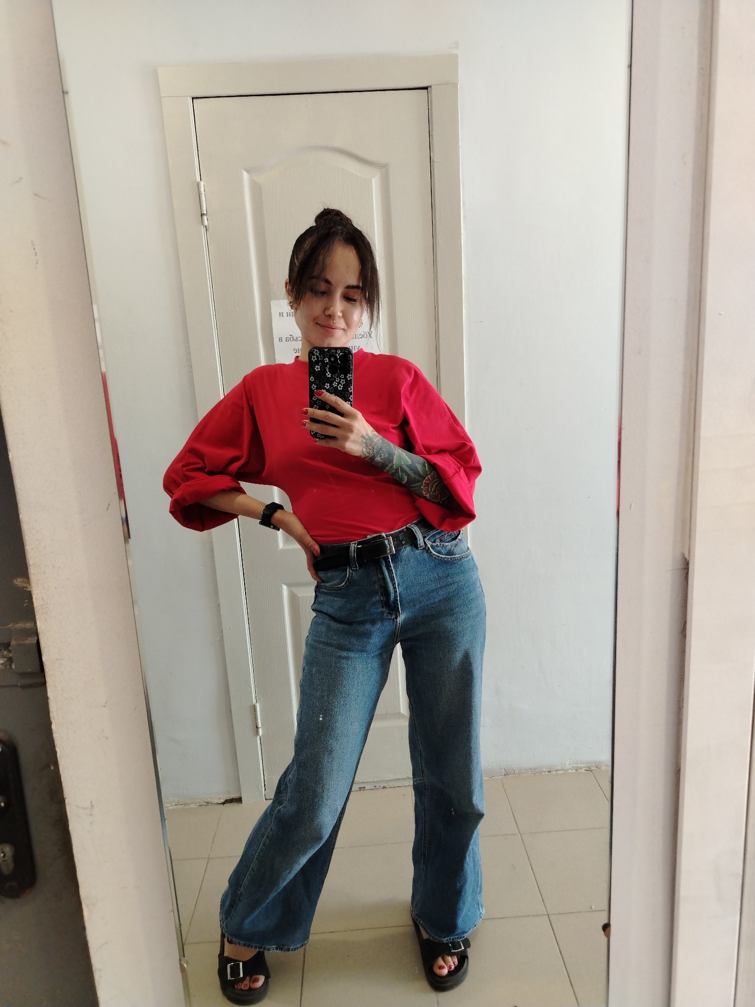 A young woman with dark hair pulled up in a bun and bangs is standing in front of a full-length mirror, taking a selfie with her phone. She is wearing a vibrant red long-sleeved shirt with voluminous sleeves and blue denim jeans. The jeans appear to be a wide-leg or flare style. She has a black belt with a silver buckle. On her feet, she wears black platform sandals with a buckle. Her left arm is bent at the elbow with her hand on her hip, and her right arm is extended to hold the phone. She has tattoos on her left arm. The background appears to be an interior space with a white door and white walls. There is a tiled floor. The image was taken in the city of Chaikovsky, Russia.