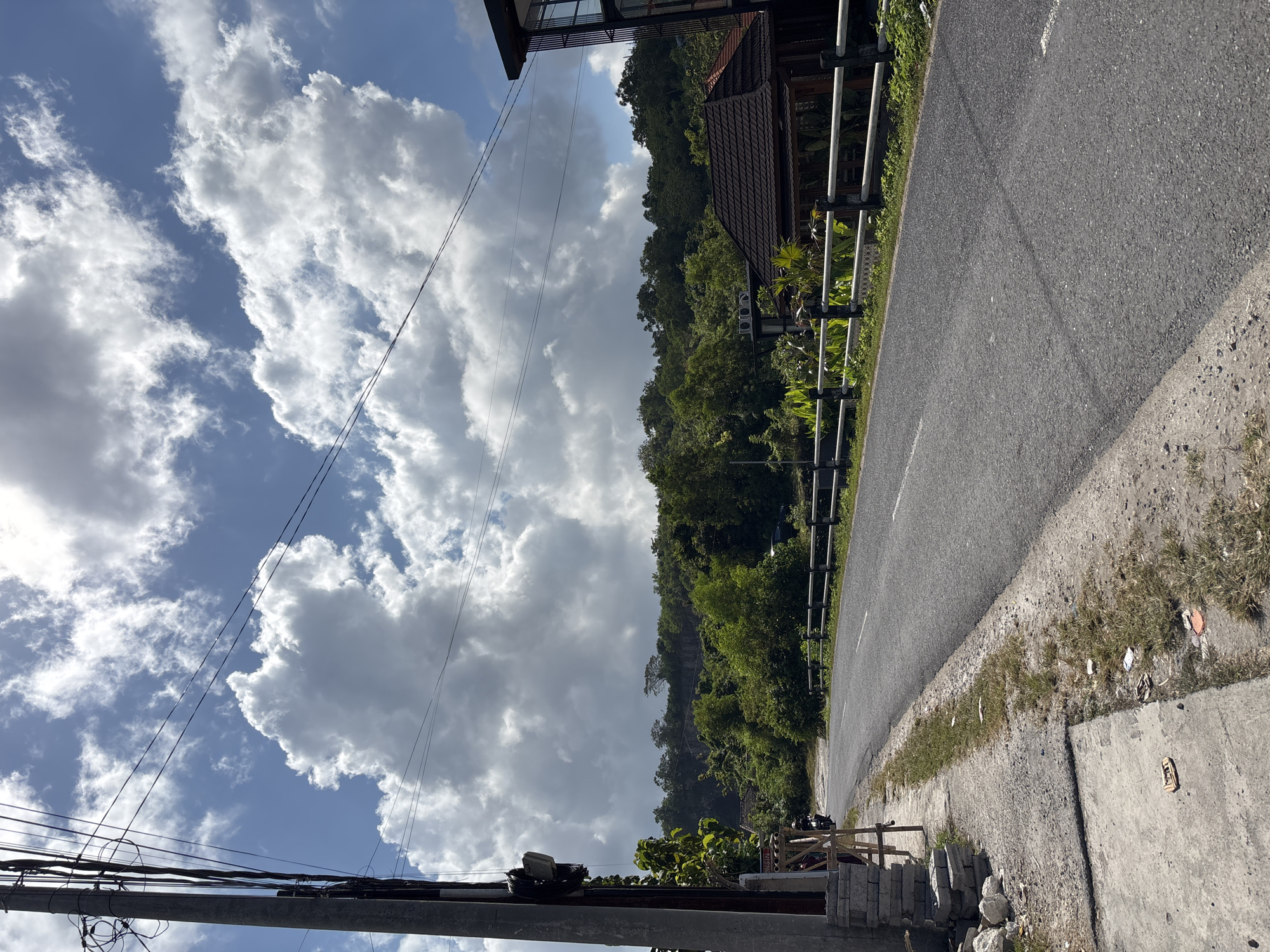 The image, when properly oriented, captures a vibrant outdoor scene featuring a paved road winding alongside a lush, green hillside under a bright, cloudy sky. The perspective is from a roadside vantage point.
In the foreground, a dark utility pole with a dense network of overhead power lines and cables occupies the lower left corner. Above these lines, a clear blue sky is dotted with large, fluffy white cumulus clouds, suggesting a sunny day.
An asphalt road, with faint white markings, stretches from the bottom right towards the upper left of the frame. It is bordered on its immediate right by a strip of dry grass and dirt, which then meets a concrete pavement or ground area, where some stacked concrete blocks and scattered debris are visible. On the left side of the road, a metal railing or fence runs parallel to it.
Beyond the railing, a steep, densely forested hillside rises dramatically, covered in rich green tropical foliage that dominates the mid-ground. Partially nestled within these trees is a building with a distinct brown tiled roof, likely a house or guesthouse. Another building structure is partially visible at the far upper left, integrated into the natural landscape.
The scene conveys a serene, semi-rural environment, characteristic of a tropical region with abundant vegetation. The strong sunlight indicates it is daytime. No people or vehicles are present, and there is no visible text in the image. The setting aligns with the provided location context of Kuta Selatan, Indonesia.