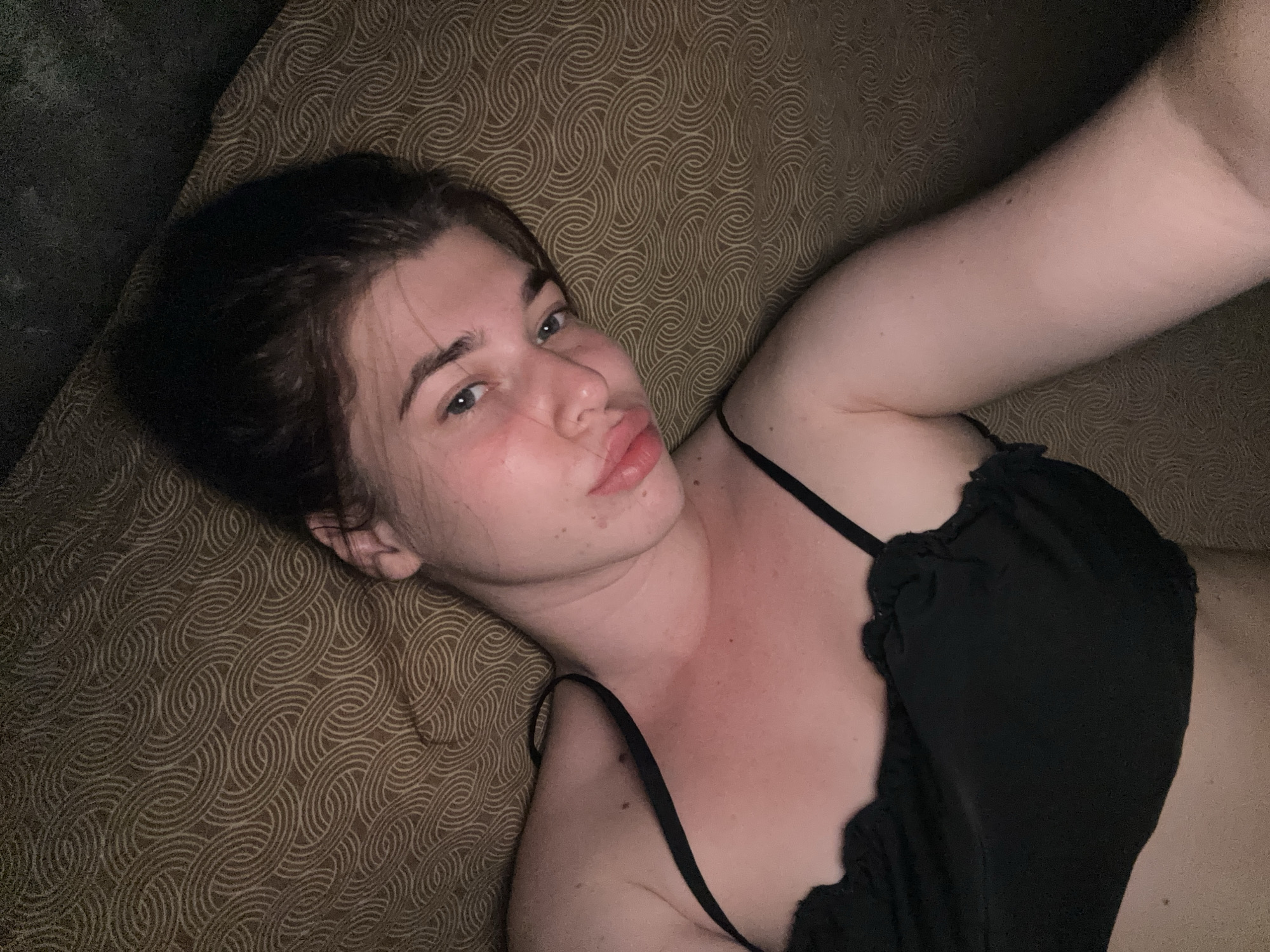 A young person with dark hair is lying on their back on a textured brown surface. They are wearing a black garment with thin straps. The person's head is tilted slightly to the side, and their gaze is directed towards the camera. Their lips are slightly parted, giving the impression of a pensive or playful expression. The lighting suggests an indoor setting, possibly at night or in a dimly lit room. The patterned background fills the frame, creating a somewhat intimate and cozy atmosphere. No text is visible in the image.