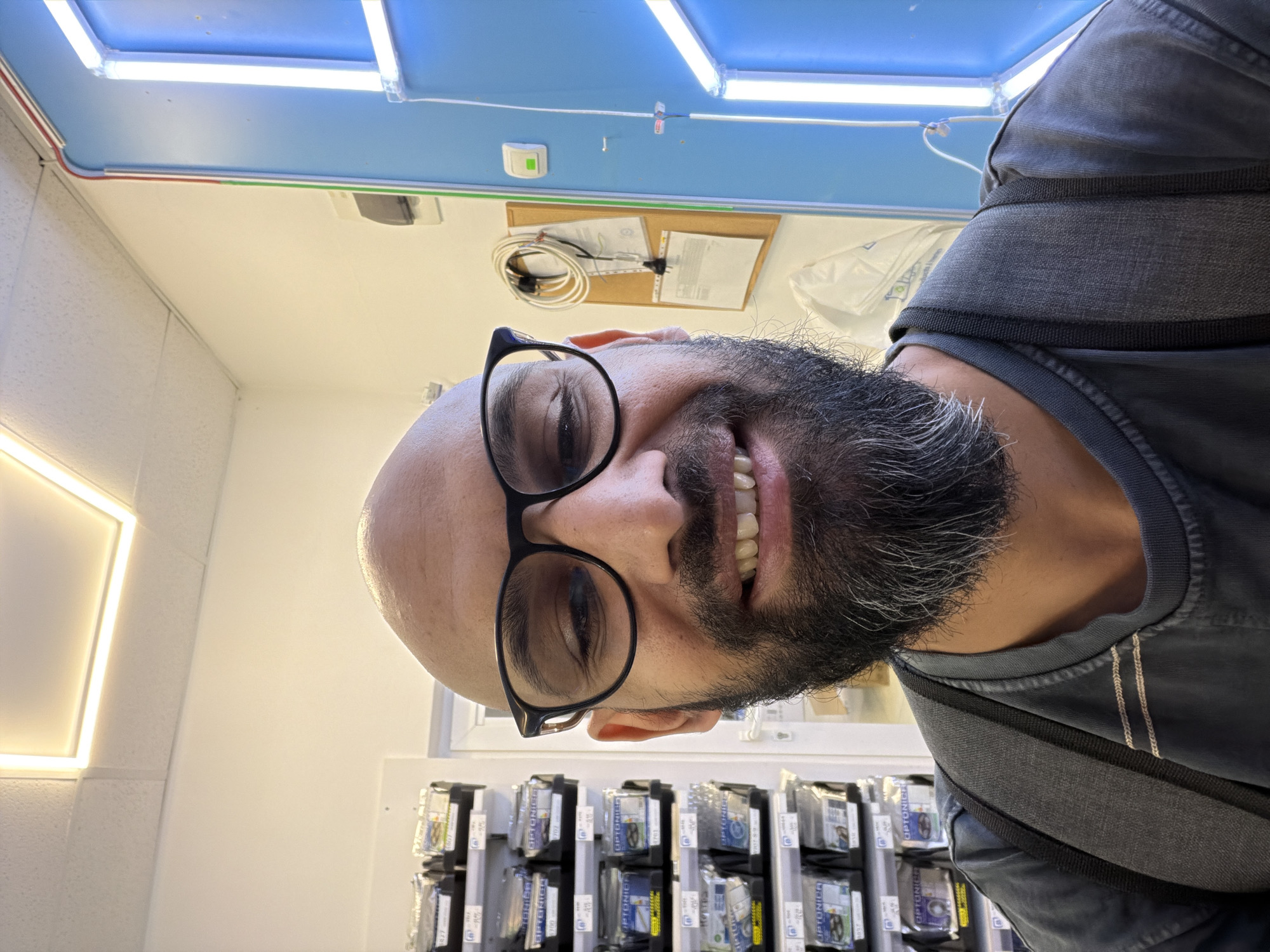 A bald man with a beard and glasses smiles for a selfie. He is wearing a dark t-shirt and a grey backpack. Behind him are shelves stocked with small, packaged items, a corkboard with papers and cables, a blue door with a light fixture, and a white ceiling with a square light panel. The context of the scene suggests it may be a retail or storage environment, potentially within a commercial establishment. The specific location in Plovdiv, Bulgaria is not discernible from the image alone.