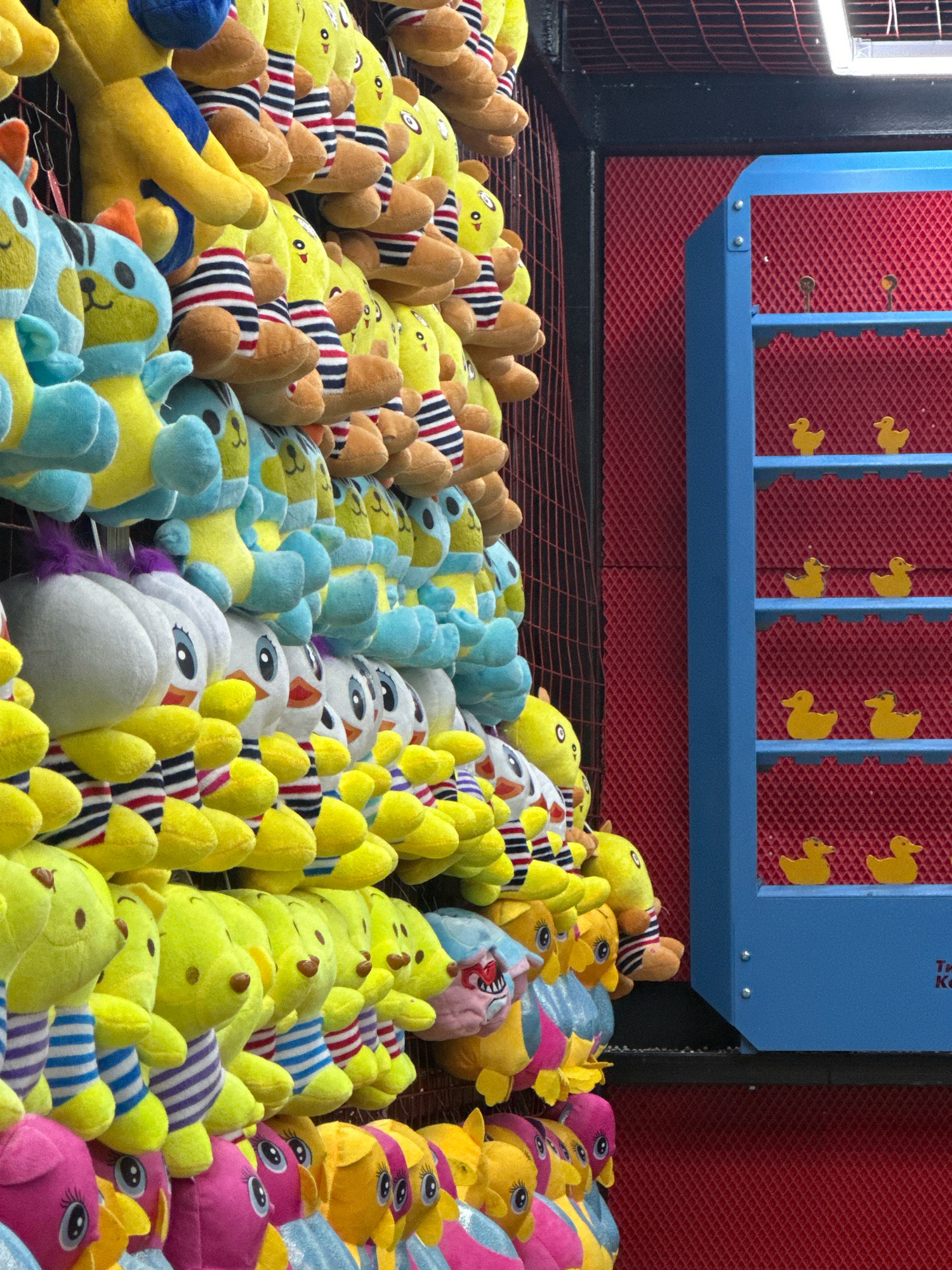 The image depicts an arcade game stall filled with plush toys. The toys are arranged in rows and are predominantly yellow, blue, and white, with some featuring striped clothing. To the right of the plush toys, a blue structure with multiple shelves is visible. Each shelf contains small, yellow duck figures. The background wall is a red diamond-patterned material. The scene appears to be outdoors, possibly at a fair or amusement park. There are no people or animals visible in the image. The image is taken from a side angle, focusing on the abundance of stuffed animals and the duck game.