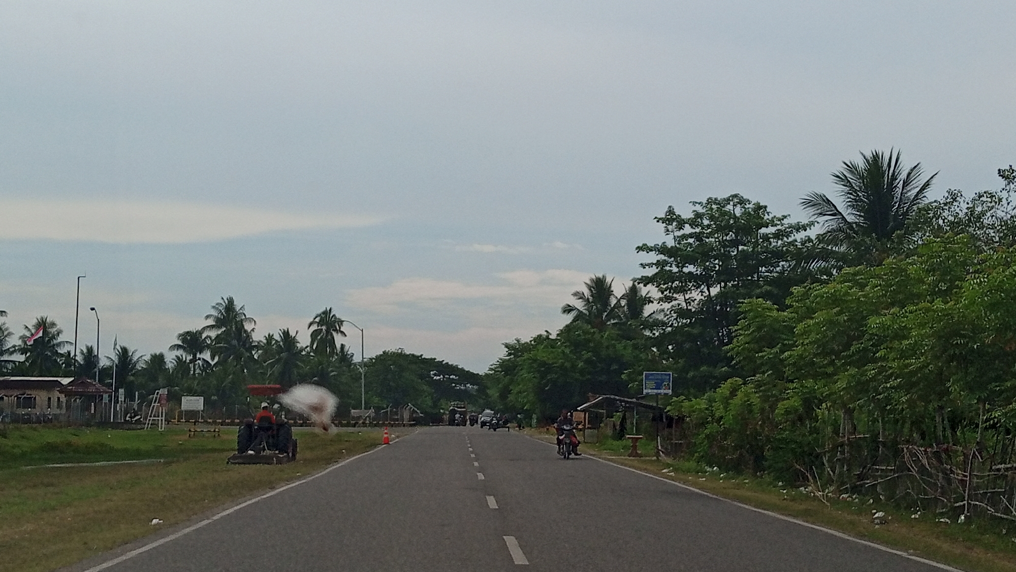 The image depicts a rural road in Glokaron, Indonesia, with lush green vegetation and palm trees lining both sides. The sky is overcast with soft clouds.
On the left side of the road, there is a grassy area with a small building and an Indonesian flag flying from a pole. A tractor is visible in the foreground, with a person operating it, and a white mist is being sprayed from it, likely for irrigation or pest control. Several other structures and what appear to be temporary barriers are also present in this area.
The road itself is paved and has a dashed white line marking the center. Several vehicles are on the road, including motorcycles with riders. On the right side of the road, more dense green foliage and trees are visible, with a small, open-sided shelter.
The overall impression is one of a working rural landscape on a cloudy day.