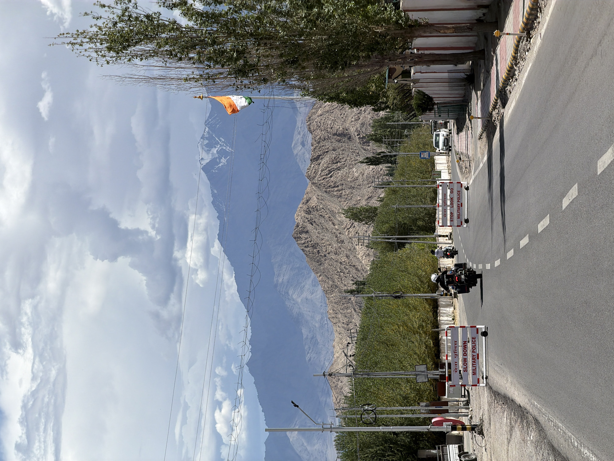 The image depicts a scenic view of a road in the city of Leh, India, with the Himalayas in the background. The road is paved and has white dashed lines marking the lanes. There are several military police barriers on the side of the road, with "SLOW DOWN" and "MILITARY POLICE" written on them in English and Hindi. Two motorcycles are visible on the road, one in the foreground and another further ahead. A white car is also seen in the distance. The sky is cloudy with patches of blue, and tall trees line the right side of the road. An Indian flag is flying on a flagpole in the distance, against the backdrop of snow-capped mountains. The environment appears to be a well-maintained road within a mountainous region, likely a security checkpoint or a control zone due to the presence of military police.