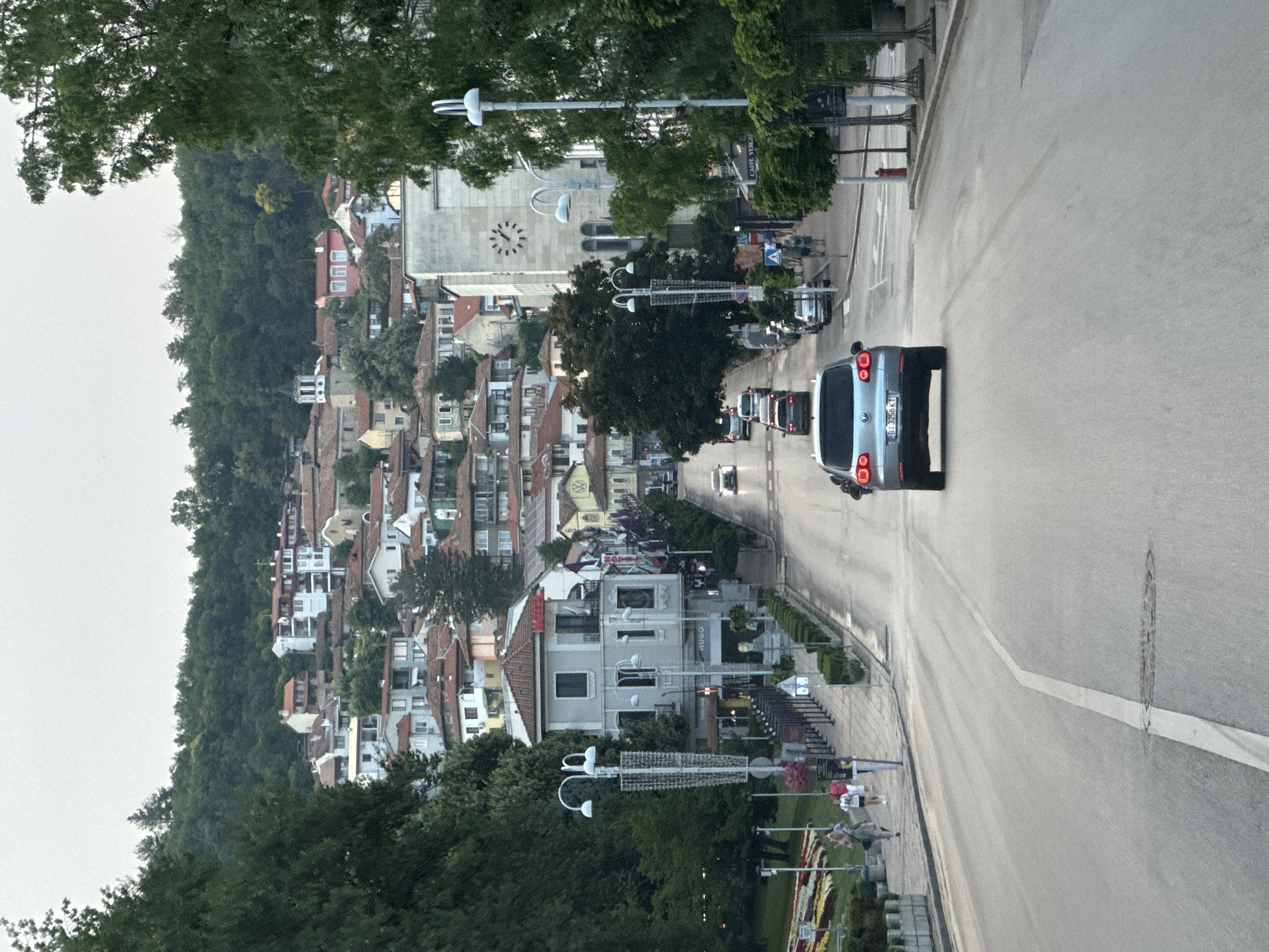 The image depicts a street scene in Veliko Tarnovo, Bulgaria, characterized by a hilly landscape dotted with traditional architecture. A paved road runs along the right side of the frame, with a silver minivan prominently parked on it. The town is built on a steep incline, with numerous buildings featuring terracotta roofs and white or pastel-colored facades cascading down the hillside. A large church with a clock tower stands out against the urban sprawl. Lush green trees frame the scene, particularly on the left and in the foreground. Several streetlights are visible, some with modern designs. A few people are walking along the sidewalk on the left side of the road. The sky appears overcast, suggesting a cloudy day. The overall impression is of a historic European town with a vibrant street life. Visible text includes "CAFE VERG" on a sign and "HUGO" on a building. The license plate of the silver minivan shows "BG 3256 KT".