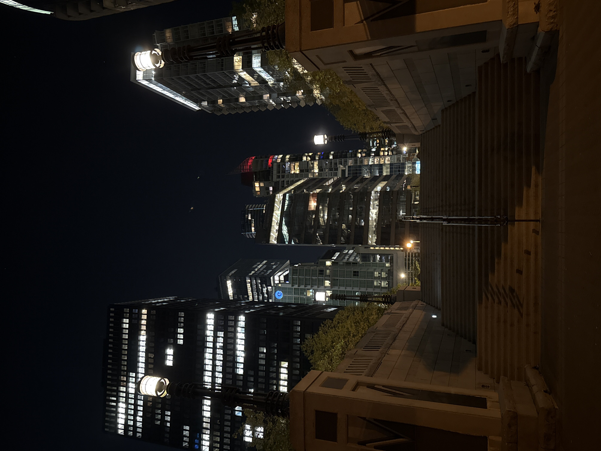 At night in Vancouver, Canada, a view looks upwards at a set of buildings from street level. The perspective is upwards, showing a stepped stone structure on the right side, leading to the sky which reveals a set of illuminated buildings, mostly skyscrapers with many windows showing light. Street lamps are present along the steps. The dark sky occupies a large portion of the image, contrasting with the buildings' lights. The buildings appear contemporary, with glass and steel construction.