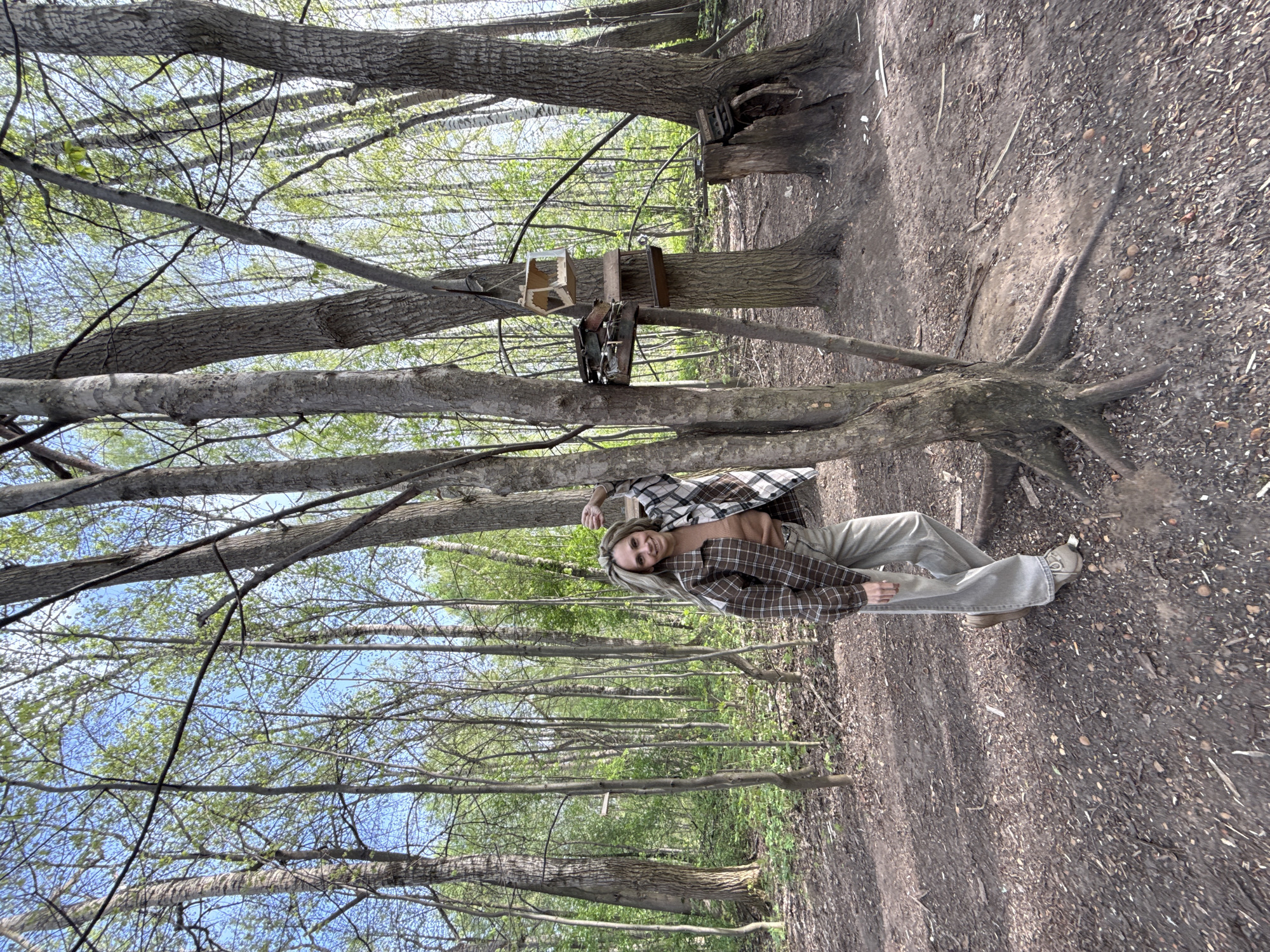 A vertical shot shows a woman leaning against a tree in a wooded area. The woman is smiling at the camera. She has blond hair and wears a brown sweater, a brown and white plaid jacket, tan pants, and tan shoes. Several trees are in the background. Some have birdhouses attached to them. The ground is dirt and leaves. The scene is in Rusanovo, Russia.
