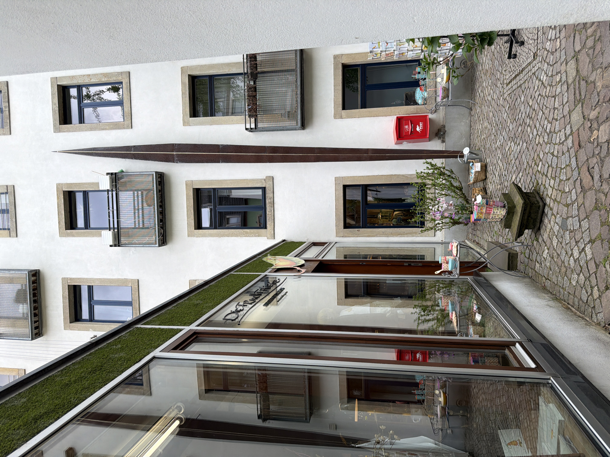 The image shows the exterior of a building. The building has a white facade with several square windows outlined with tan-colored borders. Some windows have metal barriers outside. A long, narrow, dark-colored metal object is mounted horizontally on the wall, between rows of windows. A red mailbox is visible on the wall near one of the windows. The ground is paved with cobblestones, and there's a decorative plant arrangement in a basket near the building's entrance. The building's entrance is characterized by large glass windows and doors, with some green foliage on the top of the building's overhang. The name "Die Truheide" is visible on the building's signage. The location is in the city of Unknown, Unknown.