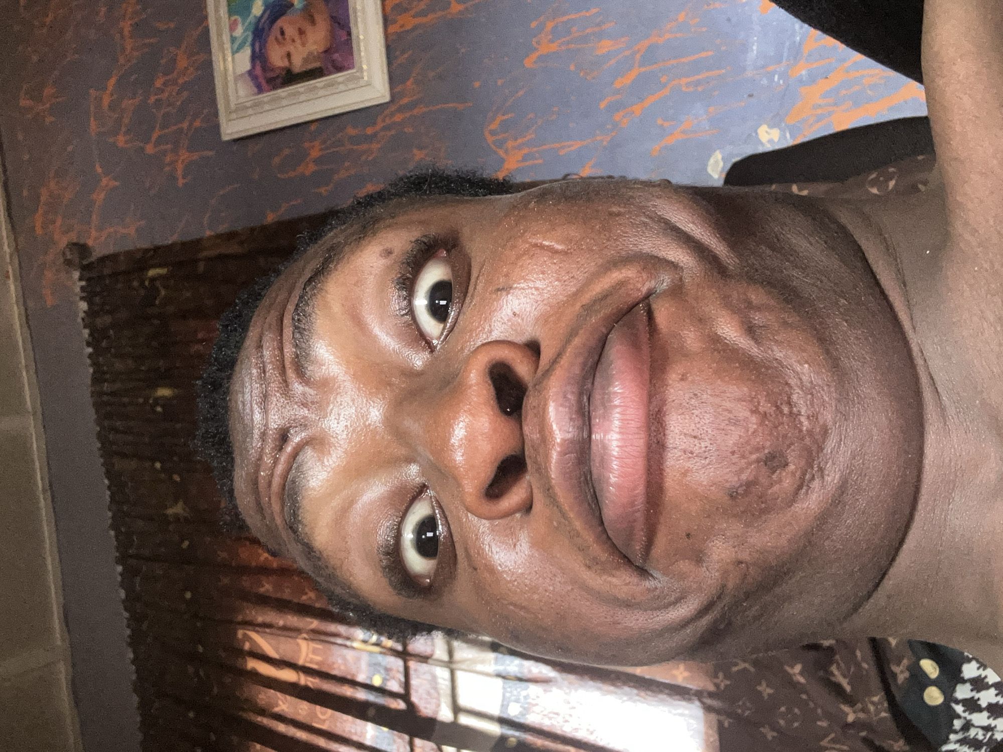 A close-up selfie of a dark-skinned person with brown eyes. The background shows a wall painted with orange and gray patterns, a framed picture, and a window with curtains. The location is not specified.