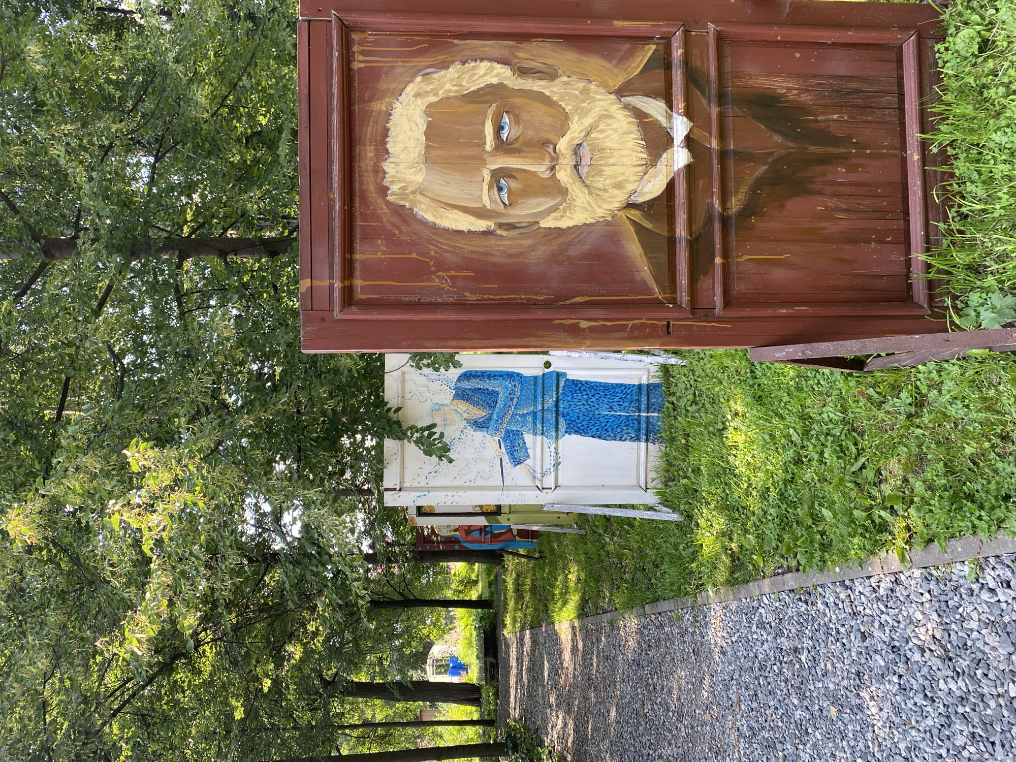 An outdoor art installation features painted doors set up in a grassy area next to a gravel path in Votkinsk, Russia. The closest door, painted a dark red, displays a portrait of a man with blond hair and a beard, intense blue eyes, and a stern expression, wearing a dark suit and white shirt with a bow tie. Behind this door, a white door showcases a painting of a figure in a blue garment, possibly holding a baton. Further down the path, other painted doors and objects are visible, partially obscured by trees. The environment consists of lush green grass, mature trees with dense foliage, and a gravel walkway. The overall scene suggests an outdoor exhibition or a public art project in a park-like setting.