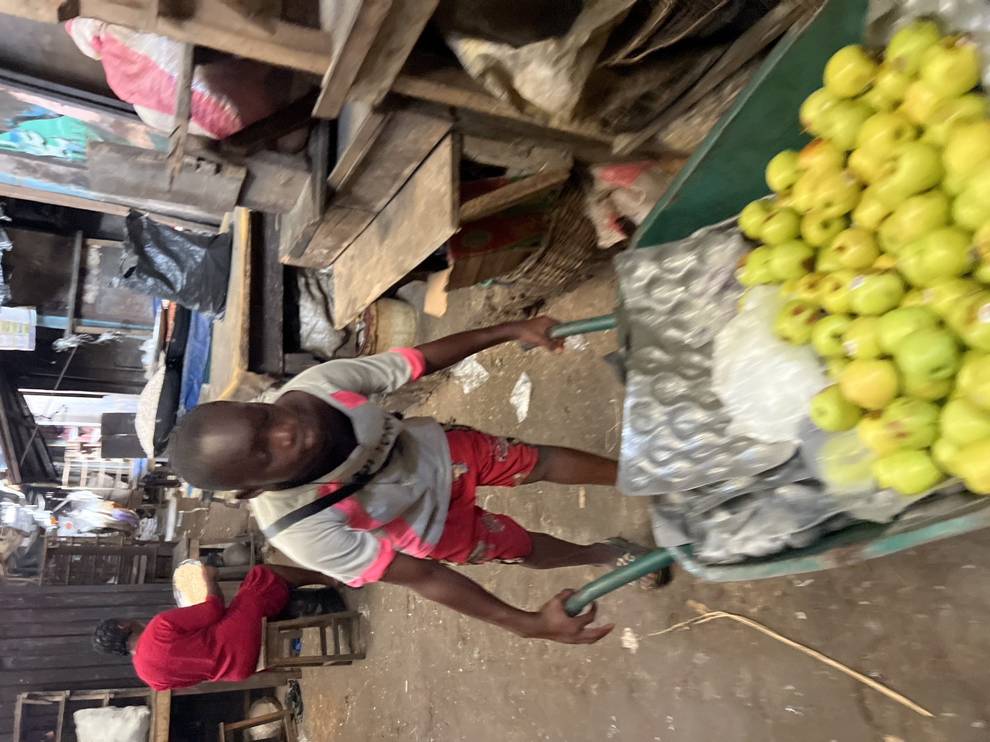 In a bustling open-air market in Nkpor, Nigeria, a man pulls a green wheelbarrow loaded with green apples, some of which are wrapped in clear plastic. The ground is concrete and somewhat dirty. The market is crowded, with wooden stalls and people visible in the background. A seated woman in a red outfit is visible in the bottom left corner. The lighting suggests it is daytime.
