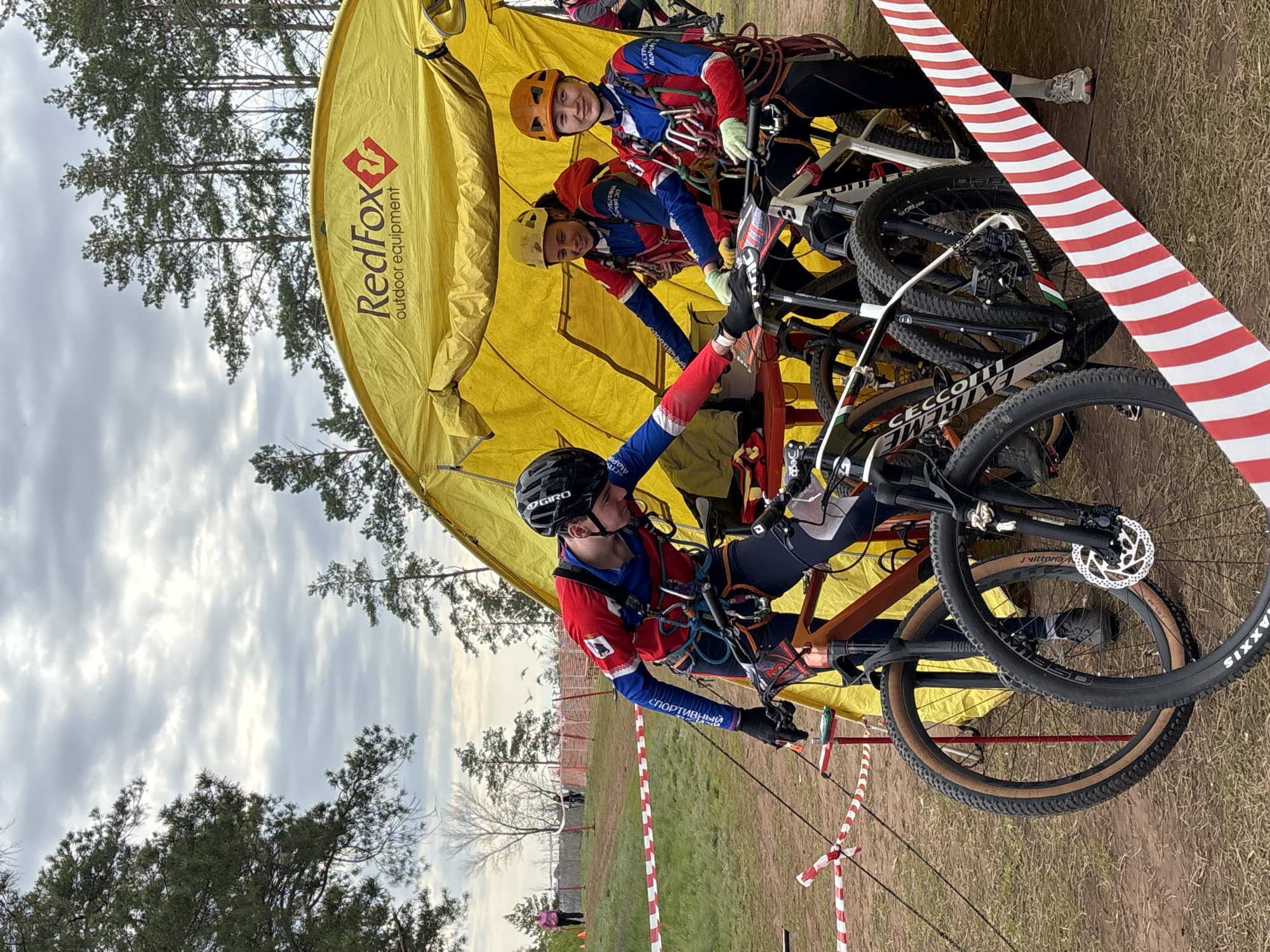 The image depicts an outdoor event in Orenburg, Russia, possibly a climbing or cycling competition. A yellow tent with the "Red Fox outdoor equipment" logo stands at the edge of a grassy area. Three people wearing helmets and matching red, blue, and white athletic gear are present. One individual is in the foreground, secured with climbing ropes and attached to a bicycle. The other two are behind, interacting with other bicycles. Several mountain bikes are clustered near the tent. Red and white striped tape forms a barrier. Trees and cloudy sky are visible in the background. The ground appears to be a mix of grass and dirt.
