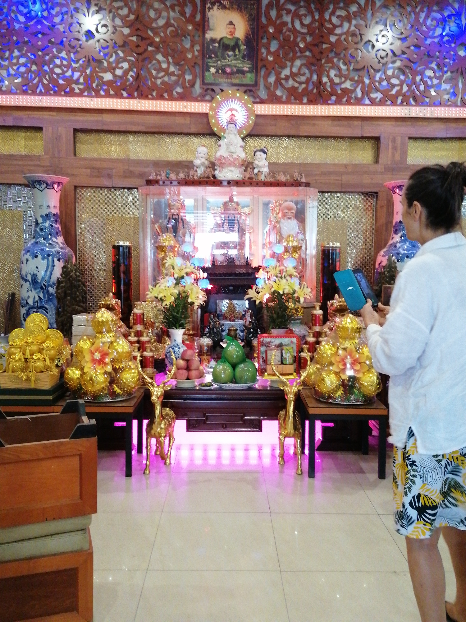 The image depicts an indoor scene in Chu Văn An Ward, Vietnam, likely within a religious or cultural setting. A woman, wearing a white shirt and patterned shorts, is holding a smartphone and appears to be taking a photograph of an elaborate altar. The altar is adorned with golden statuettes, flower arrangements, and displays of fruit. Statues of deities or religious figures are visible in the background, behind glass enclosures. The room has ornate wooden carvings on the walls, and the floor is tiled. The lighting is a combination of natural and artificial sources, with pink LED lighting under the altar.