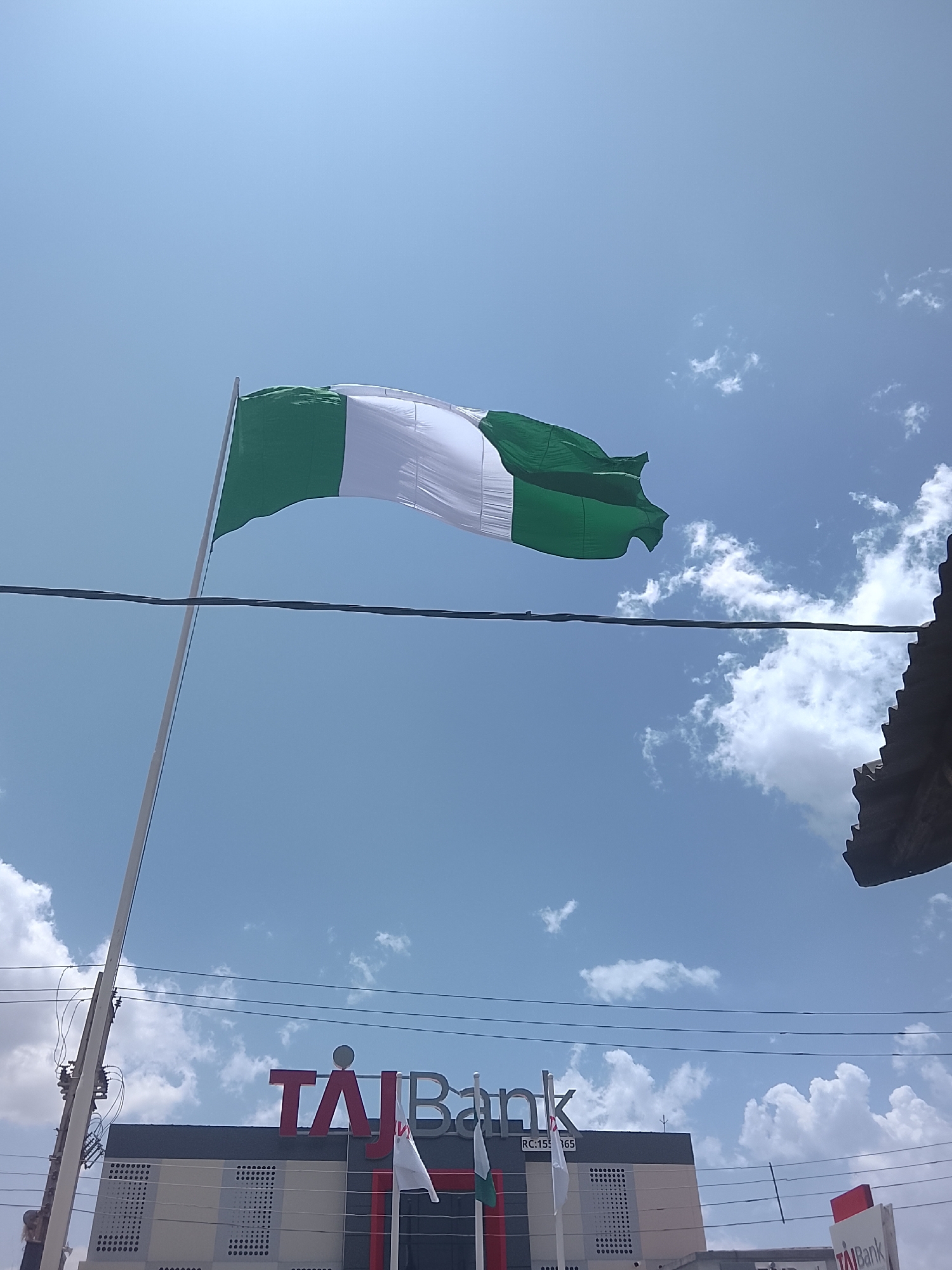 The image shows the Nigerian national flag waving in the wind against a partly cloudy sky. Below the flag is the building of TAJ Bank. The location appears to be in the city of Jos, Nigeria, due to the presence of the Nigerian flag and the TAJ Bank signage. No people are visible in the image. An electrical wire runs across the frame.