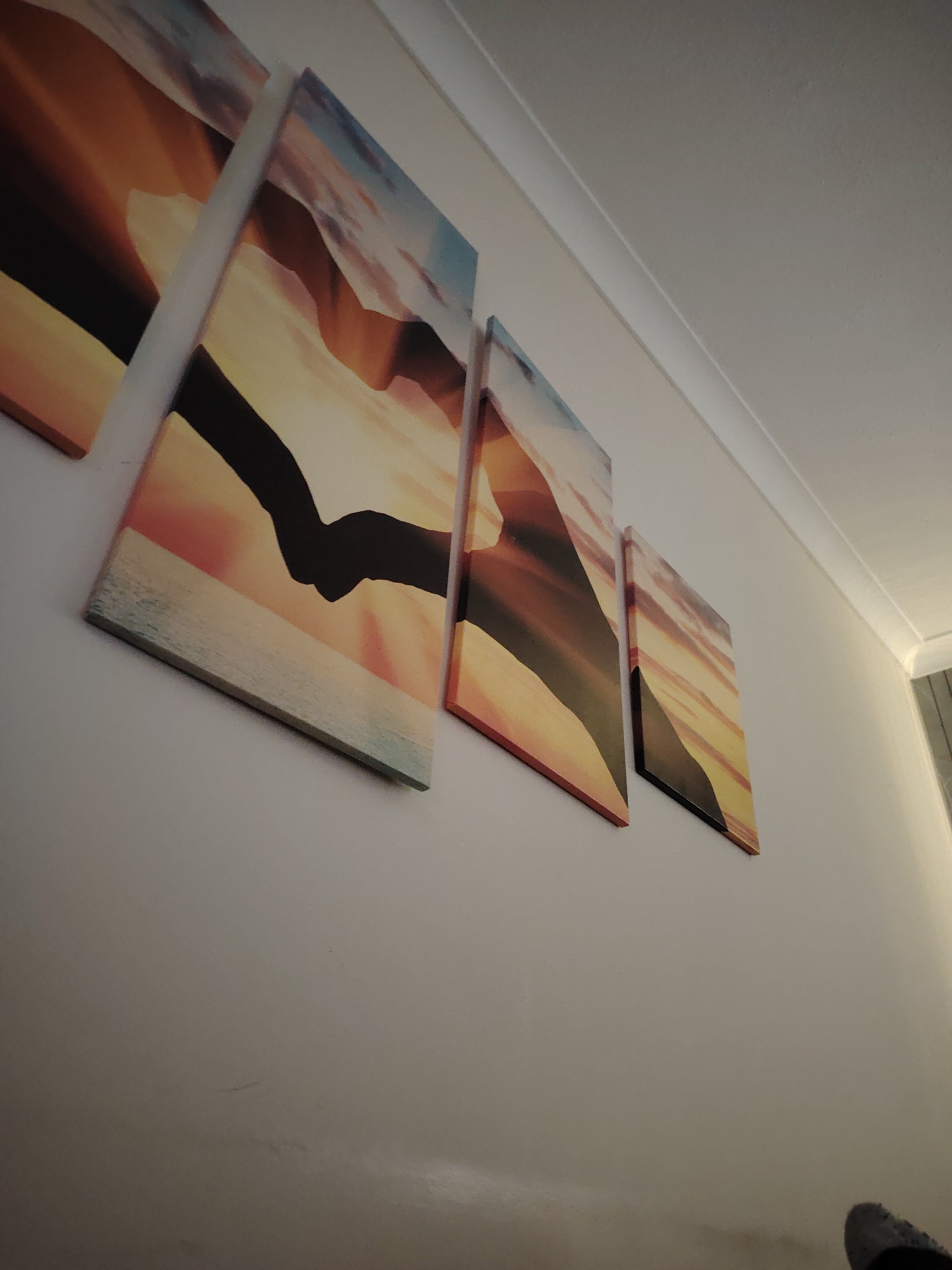 Four rectangular canvas prints are mounted horizontally on a light-colored wall in Huntingdonshire, United Kingdom. The prints, appearing to decrease in size from left to right due to the upward and diagonal photographic perspective, depict stylized landscapes or abstract designs with warm hues of orange, yellow, and red, often featuring dark silhouetted forms. The second print from the left distinctly shows two hands clasped or interlocked, silhouetted against a vibrant sunset or sunrise with a visible horizon line. The wall is a uniform light color, topped by a white ceiling and decorative coving. The scene is illuminated by soft, indirect light. A dark, textured object is partially visible in the bottom right corner.