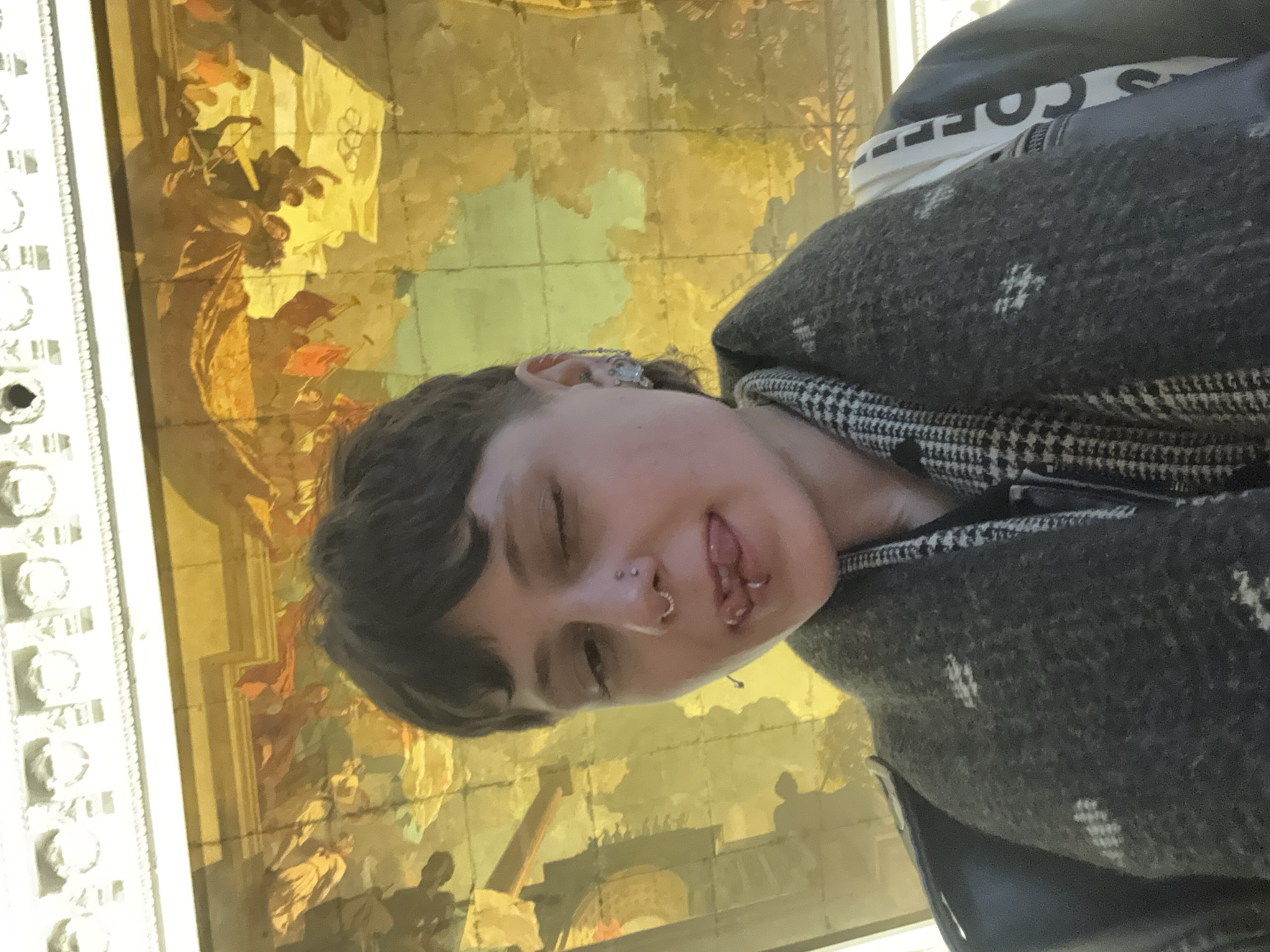 The image, presented at a 90-degree clockwise rotation, primarily features a young person looking directly at the camera, set against an elaborate, golden-hued mural. The person has short, dark hair and is captured mid-expression, with one eye winking or squinting and their tongue slightly visible, suggesting a playful or cheeky demeanor. They have several facial piercings, including a septum ring, a philtrum piercing, and potentially cheek piercings, along with an earring on their visible ear.
The individual is wearing a dark grey, textured jacket or coat, possibly wool, featuring a subtle pattern of light-colored, asterisk-like motifs. A black and white plaid pattern is visible on the garment's collar or lining. Beneath the jacket, a white layer of clothing displays partial black text, which appears to spell "COFFE" or a similar word.
The background is dominated by a large, ornate mural, likely painted on a ceiling or high wall, depicting numerous figures in what appear to be historical or mythological scenes. The mural is rich in warm, golden and earthy tones, showing architectural structures, groups of people in various poses, some holding flags or banners, and intricate detailing. Along what would be the top edge if the image were upright, a decorative white border with repeating geometric or floral patterns frames the mural.
The setting appears to be a grand interior space, such as a museum, palace, or historic building, consistent with the architectural and artistic styles found in cities like Saint Petersburg, Russia. The lighting is soft and warm, illuminating the detailed artwork.