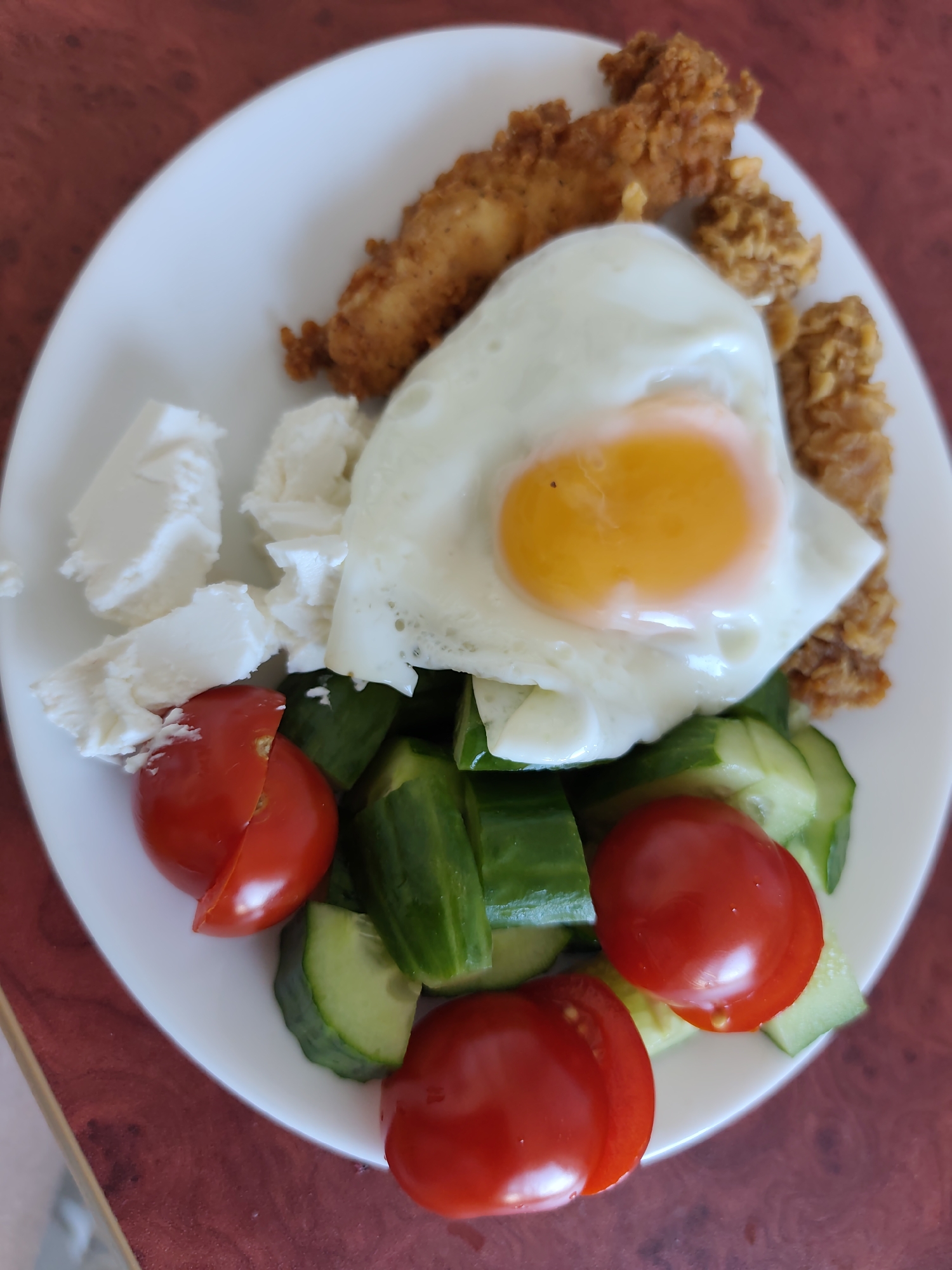 The image depicts a plate of food. The food consists of a fried egg, fried chicken, cubed white cheese, sliced cucumbers, and sliced cherry tomatoes. The plate is sitting on a table with a red-brown patterned surface. The photo was taken in Chaikovsky, Russia.