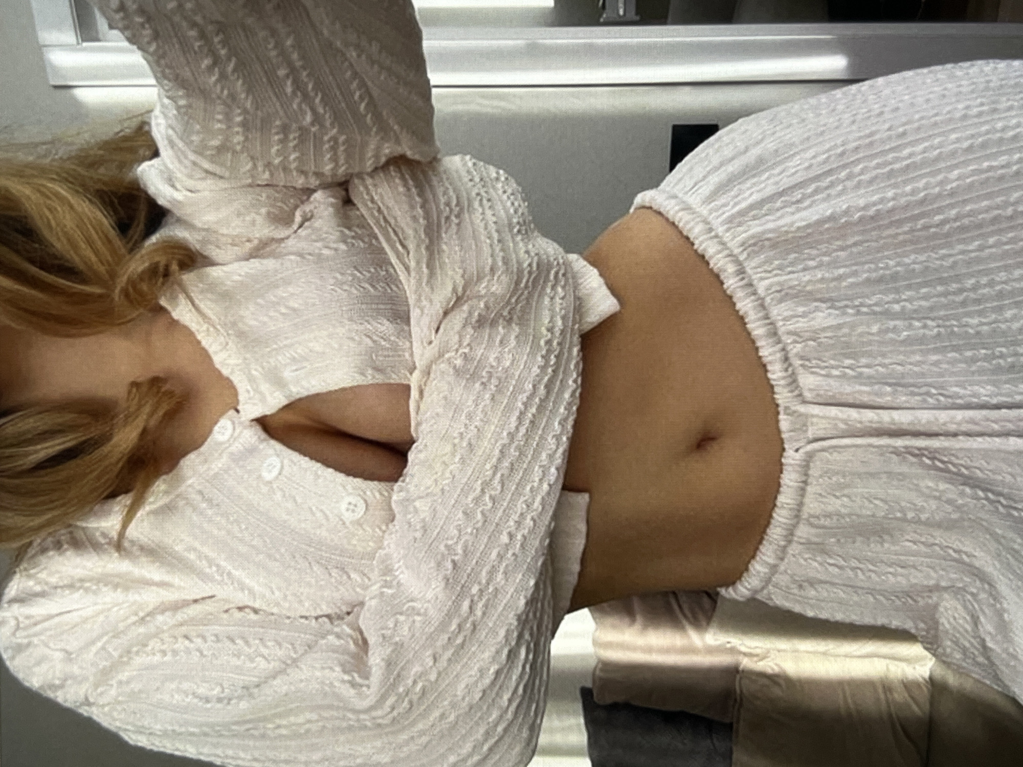 The image shows a close-up of a person wearing a white textured outfit. The top is a cropped cardigan with buttons down the front, and the bottoms are high-waisted pants with a drawstring. The person's midriff is exposed. The background is out of focus but appears to be indoors, possibly a bedroom, with soft lighting.