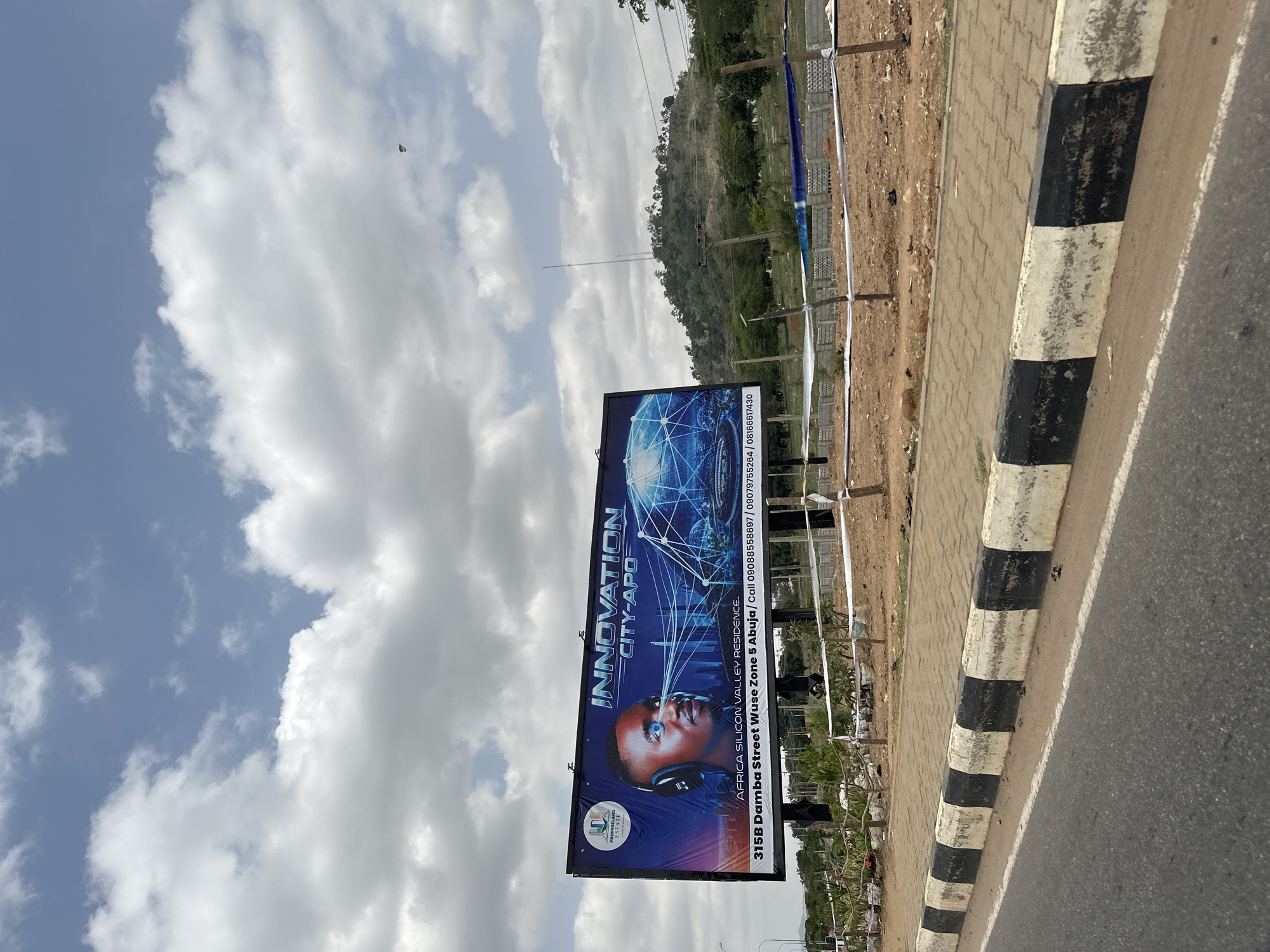 The image displays a daytime outdoor scene featuring a prominent billboard alongside a road. The foreground shows an asphalt road bordered by a black and white checkered curb. Beyond the curb, there's a paved pedestrian path that transitions into a dirt or gravel shoulder, where several small trees or shrubs are planted.
The central subject is a large advertising billboard. Its design features a stylized headshot of a person wearing headphones, with an intricate blue, networked globe overlaying a city skyline and the text "INNOVATION CITY-APO" and "AFRICA SILICON VALLEY RESIDENCE." The billboard also displays the "PROMISELAND ESTATE" logo and contact details: "315B Damba Street Wuse Zone 5 Abuja / Call 09088558697 / 09079755264 / 08166617430."
In the background, a large hill covered in green vegetation rises. A tall, thin antenna or mast stands atop this distant hill. The sky is partly cloudy, with patches of blue sky visible between large white cumulus clouds. A small, dark, indistinct object is visible high in the sky.
No specific activities are occurring, and no actual people are visible, presenting a static street scene. The bright, even lighting indicates it is mid-day. The location is confirmed as Abuja, Nigeria, based on the address provided on the billboard. The overall impression is that of an developing urban or peri-urban landscape.