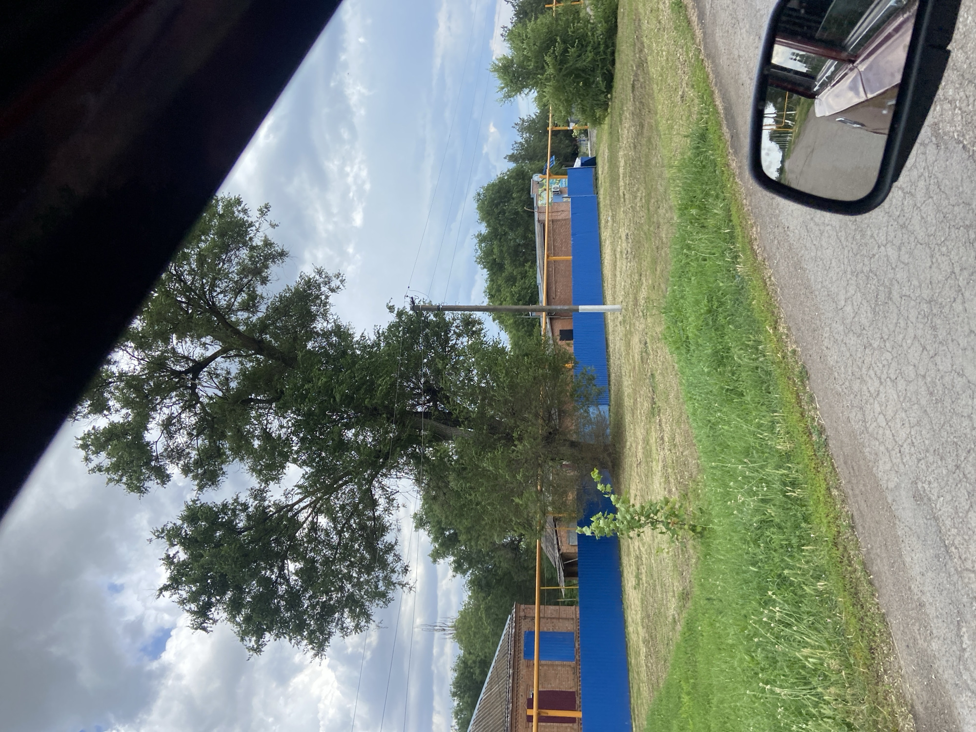 The image depicts a scene from a vehicle's side mirror, looking outwards on a cloudy day. The mirror reflects a road with a grassy verge, leading to a blue metal fence and some brick buildings. Behind the fence, trees and more buildings are visible. The sky is overcast with white clouds. The overall impression is a rural or semi-rural setting in Russia.
The primary subjects are the road, the blue fence, the brick buildings, and the surrounding vegetation. The scene is captured from a moving vehicle, as indicated by the angle of the mirror and the perspective of the road. There are no people visible, but the presence of buildings suggests human activity.
The time of day appears to be daytime, likely mid-morning or afternoon, given the lighting. The weather is cloudy, contributing to a diffused light. The location context of Taganrogsky, Russia, provides a general geographical setting.
Notable details include the bright blue color of the metal fence, contrasting with the muted tones of the brick buildings and the dry grass. The reflection in the mirror offers a glimpse of the car's interior and the passing scenery. There is no visible text in the image. The image conveys a sense of everyday travel and a view of an ordinary landscape.