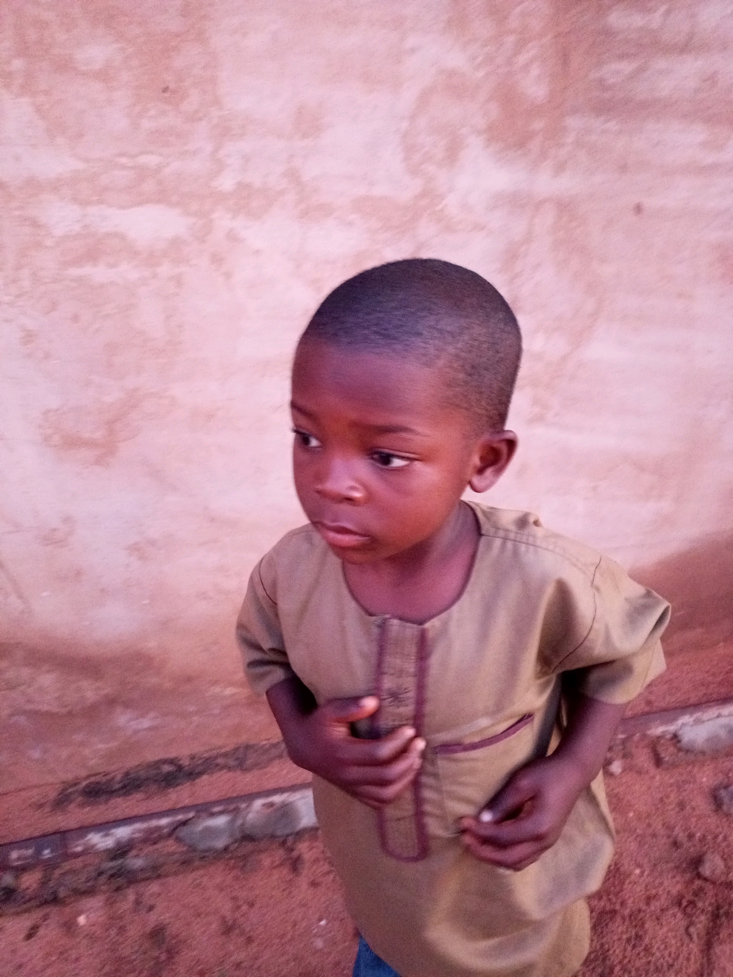 A young boy stands in front of a textured, pinkish-brown wall in Tempure, Nigeria. He is wearing a tan, long-sleeved garment with dark trim around the neckline and a decorative panel down the front. His hands are clasped in front of him. The ground beneath him is a reddish-brown, sandy soil. The boy's gaze is directed off to his left, away from the camera.