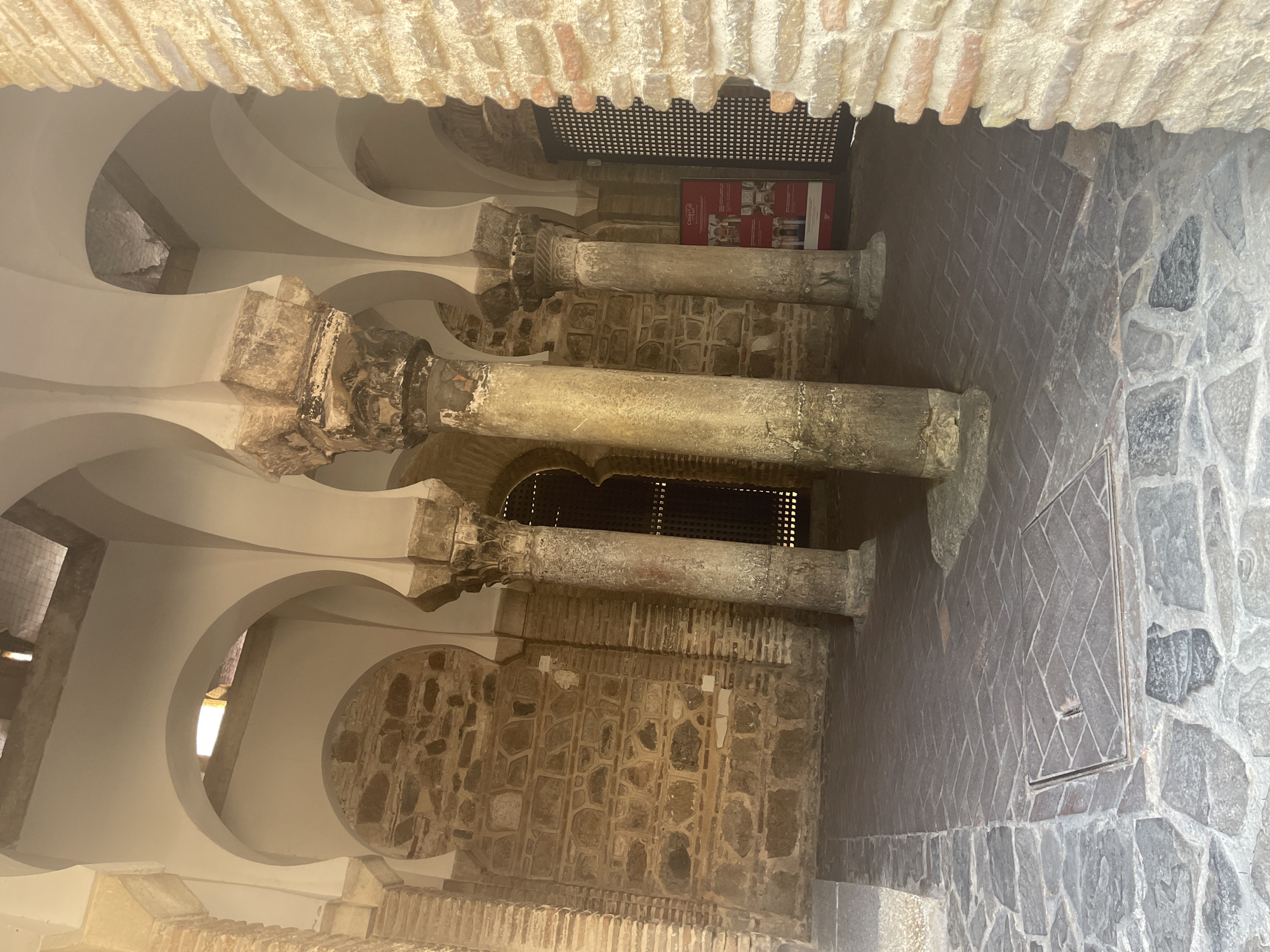 This image captures an interior view of the Mosque of Cristo de la Luz in Toledo, Spain. The scene is characterized by its distinctive Islamic architectural style, featuring a series of horseshoe arches supported by weathered stone columns. The walls are constructed from rough-hewn, multi-colored stone blocks, typical of medieval fortifications, with some areas showcasing exposed brickwork.
The floor is paved with irregular, dark gray flagstones in the foreground, transitioning to a herringbone pattern of dark bricks further back. The play of light and shadow suggests an outdoor or semi-outdoor space, likely a courtyard or ambulatory within the mosque complex, given the open arches. In the background, a dark, latticed gate is visible, hinting at further chambers or an exit.
A red informational poster with images is mounted on a pillar towards the right side of the frame, providing context about the site. No people are visible in the image. The overall impression is one of ancient architecture, preserved and displayed within the historic city of Toledo.