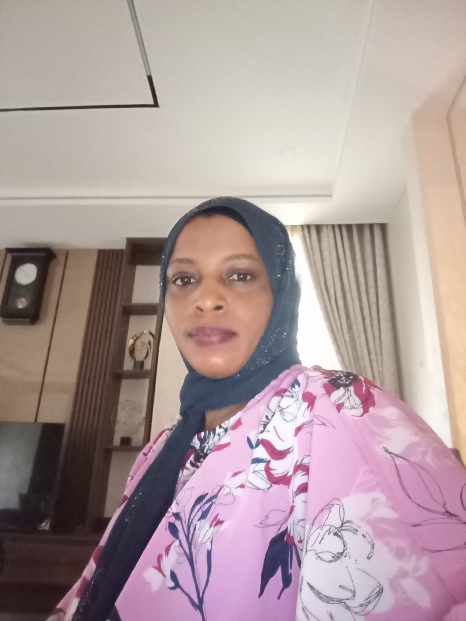 A woman wearing a dark blue hijab and a pink floral print top is posing for a picture indoors. Behind her is a wall with a clock and a wooden display case. The room has white walls and curtains. The location is not specified in the image, nor the city of Unguwar Rimi, Nigeria.