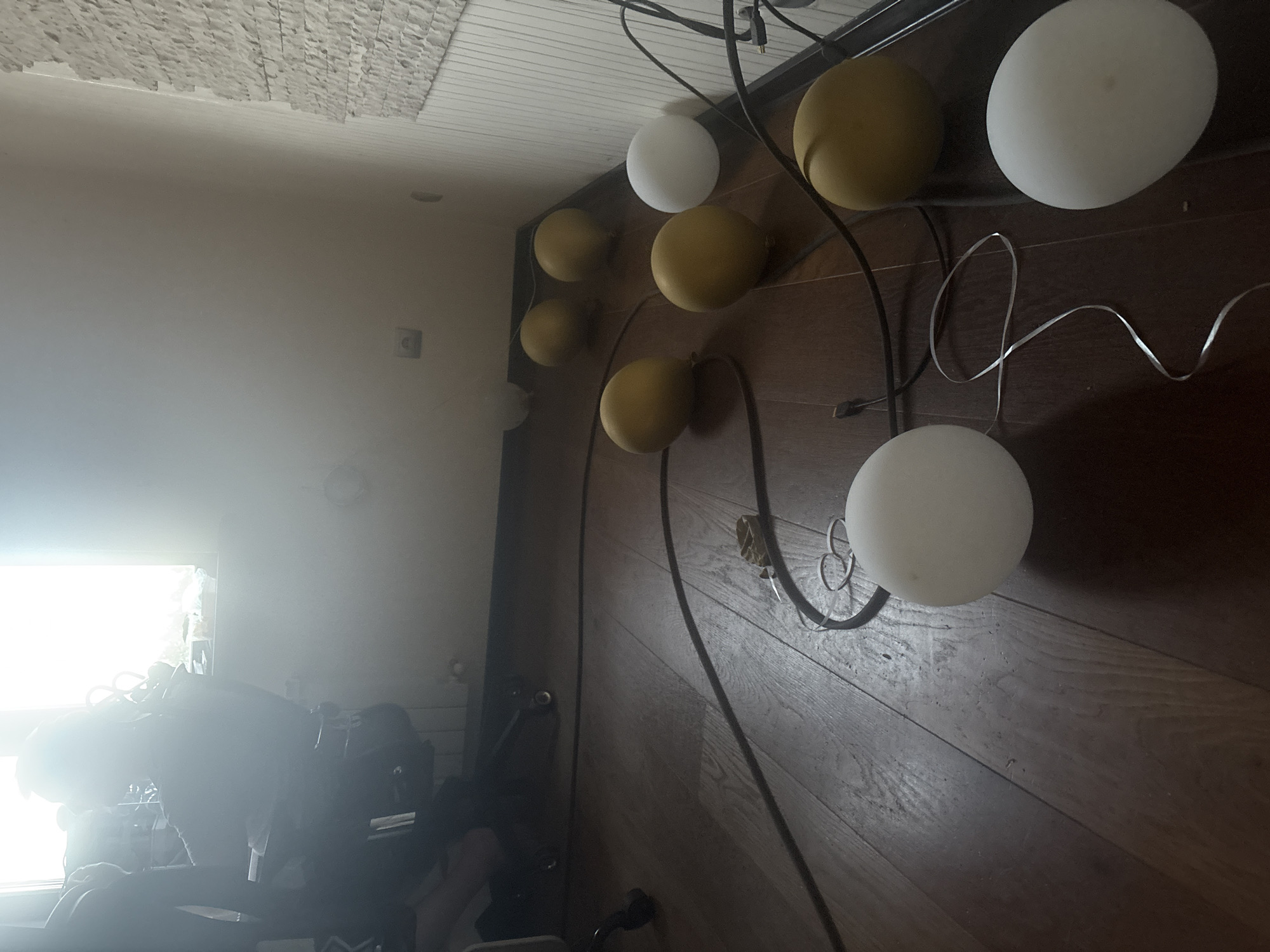The image shows a room with wooden flooring and light-colored walls. In the background, near a window with bright sunlight, a person is seated in an office chair. Several spherical lights, some yellow and some white, are scattered on the floor, connected by black cords. One white spherical light is positioned in the foreground, to the left of the frame. The wall on the right side of the image has a textured pattern. The overall impression is that of a domestic or office space with decorative lighting. There is no visual information in the image to suggest the location is in the city of Orekhovka, Russia.