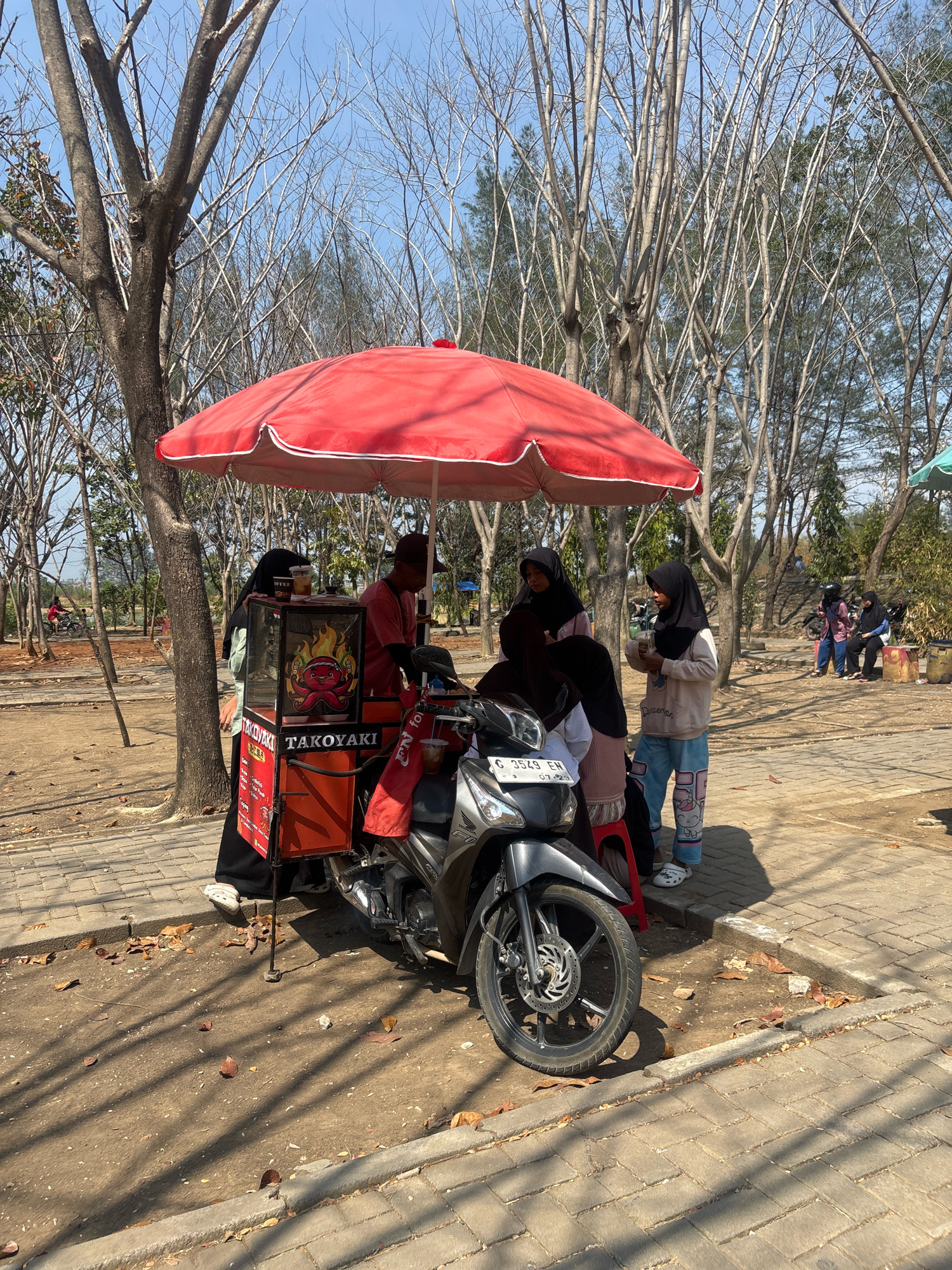 In Tegal, Indonesia, a mobile takoyaki stand operates under a large red umbrella. The stand is mounted on a motorcycle, featuring a vibrant orange and red cart displaying images of food and the word "TAKOKYAKI". A male vendor, wearing a red shirt and a dark cap, stands behind the cart, attending to customers. Several individuals are gathered around the stand, including two young women in hijabs and light-colored tops, one of whom is holding a drink. A third young woman, also in a hijab and a light pink shirt, stands near the cart. In the background, other people are visible in the distance, some sitting on benches or riding motorcycles. The environment appears to be an outdoor area with dry, leafless trees casting long shadows on the paved ground and dirt paths. The sky is clear and blue.