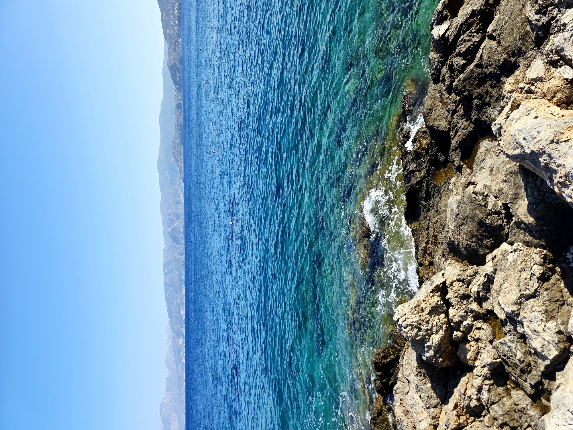 This is a landscape photograph taken from a high vantage point overlooking a rocky coastline with the sea. The image is oriented vertically, with the sky on the left and the sea and rocks on the right.
The sea is a vibrant turquoise and deep blue, with gentle waves lapping against the shore. The water is exceptionally clear, allowing glimpses of the rocky seabed beneath. The coastline is characterized by rugged, sun-baked rocks of varying sizes and textures. Some of these rocks are covered in sparse vegetation. White foam from the waves breaks against the rocks, creating a dynamic contrast with the stillness of the water and land.
In the distance, across the sea, a hazy, mountainous landscape can be seen. The slopes of the mountains are covered in a light green or greyish-brown hue, suggesting a dry, Mediterranean climate. The sky is a clear, bright blue with no visible clouds, indicating a sunny day.
A single, small figure, possibly a person swimming or floating, is visible in the expanse of the blue sea, providing a sense of scale to the vastness of the water. The time of day appears to be midday or early afternoon, given the bright sunlight and the shadows cast on the rocks.
The overall impression is one of natural beauty and tranquility. The scene evokes a sense of peace and serenity, typical of a coastal vacation destination in Greece, specifically Agios Nikolaos Municipal Unit, Greece. There is no visible text in the image.