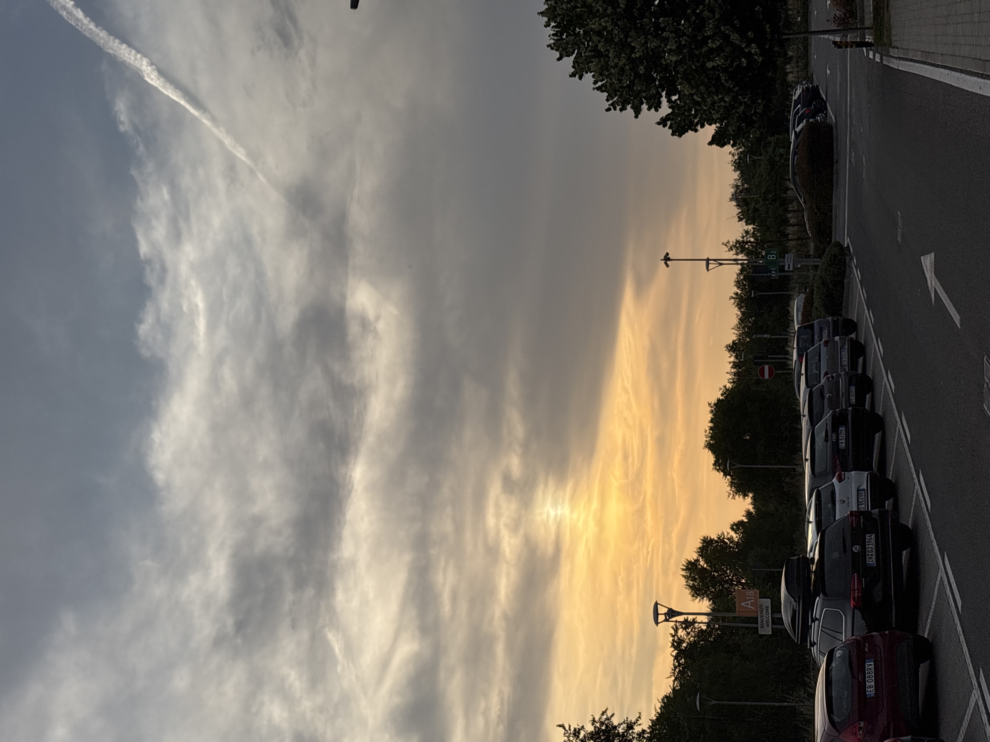 The image captures an outdoor scene at either sunrise or sunset, characterized by a dramatic sky transitioning from soft grey-blue on the left to vibrant orange and yellow hues on the right, indicative of the golden hour. A distinct, white contrail from an aircraft is visible stretching diagonally across the upper left portion of the sky.
In the foreground and midground, an asphalt road with white painted markings, including a directional arrow and parking bay lines, is visible. A line of parked cars is arranged along the right side of the road. Notable license plates include "EB-088RV" on a red car, "IN D3 706A" on a grey SUV, "D 864VR" on a white car, and "FB 843MK" on a black car. A partial view of other cars is also present.
Along the left side of the road, a dense line of dark green trees and bushes forms a boundary. Several lampposts are interspersed among the trees and along the roadside. A tall, multi-panel sign stands prominently, displaying "A18" at the top, followed by "BENVENUTI" and "WELCOME" in white text on a dark background. This suggests an entry or welcome point, potentially for a highway or a large facility in Rome, Italy, given the context.
The scene conveys a tranquil atmosphere, with the striking colors of the sky being a central visual element. The weather appears fair, with scattered clouds.