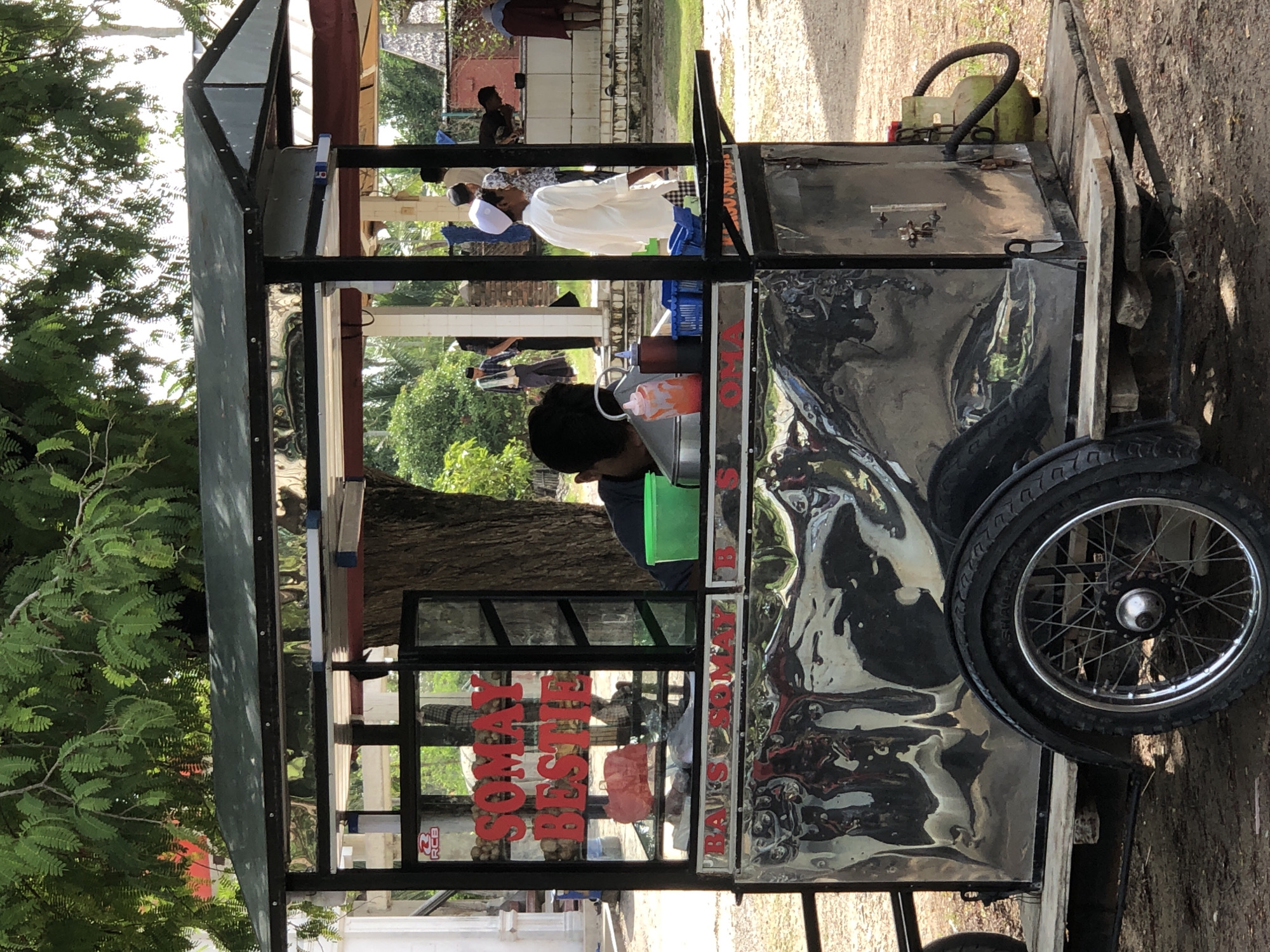 This is a medium shot, eye-level, outdoor photograph of a somay cart parked in what appears to be a courtyard or open area, likely in Palohgadeng, Indonesia, based on the accompanying information. The cart, made of metal and wood, has a dark green canopy. On the front of the cart, red lettering spells out "BAIS SOMAY B S". The cart is adorned with reflective chrome panels that show distorted reflections of the surroundings.
A person, partially visible from the back, is seen behind the counter, wearing a dark shirt. In front of them, on the counter, is a bright green plastic container and a silver pot. There are also two condiment bottles, one orange and one dark red, and a blue plastic basket. A young person wearing a white shirt and a white cap stands to the right of the cart, looking away from the camera.
In the background, several other people are visible. Some are standing near a tiled wall, and one person appears to be bending down. The environment is lush with green foliage from a large tree providing shade over the cart, and other greenery in the distance. The ground is dirt and gravel. The overall impression is a lively street scene in a tropical climate, centered around a food vendor.
