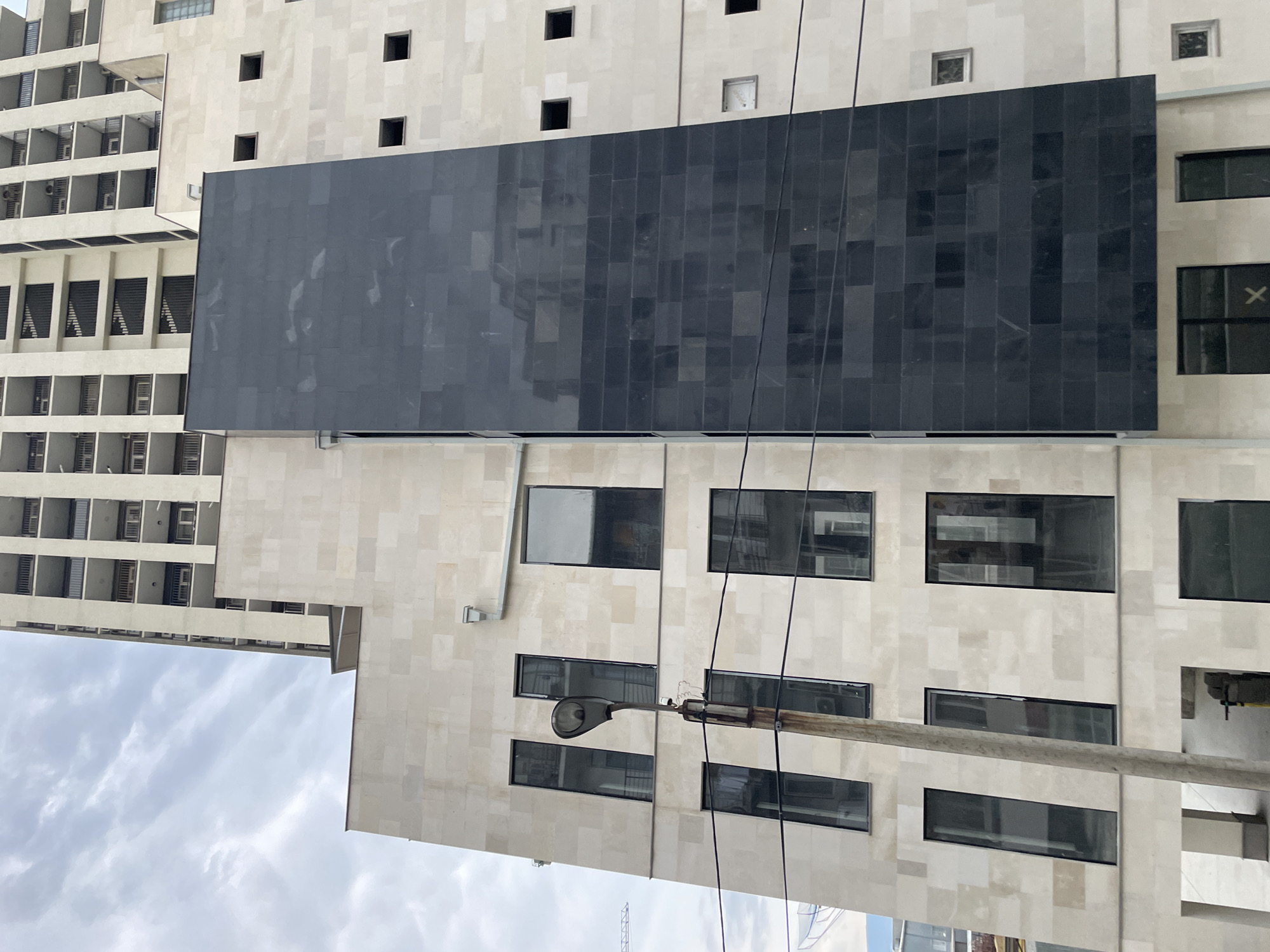 The image captures a close-up, eye-level view of a modern building in Tbilisi, Georgia. The structure features a contrasting facade, with a lower section clad in light-colored stone and a prominent, tall vertical section covered in dark, reflective tiles. Several large, dark-tinted windows are visible on the stone facade, some appearing to be floor-to-ceiling and others smaller, square openings. A weathered, rusty lamppost stands in front of the building, with wires strung across the foreground. In the background, a much taller, light-colored building with numerous balconies and stacked units rises into a cloudy sky. The overall impression is of a contemporary urban architectural scene, possibly under construction or recently completed, given the unfinished appearance of some elements and the presence of the lamppost and wires.