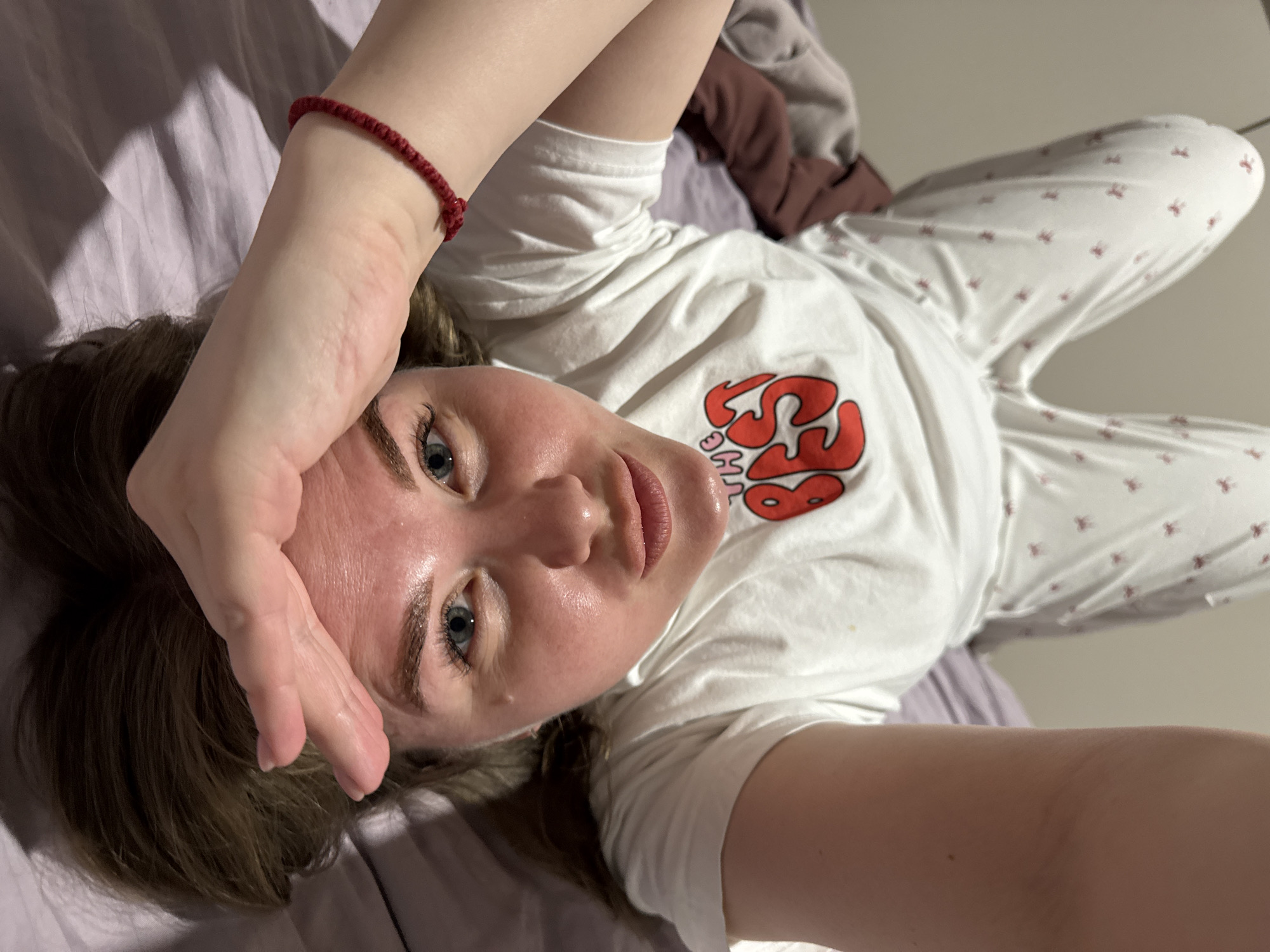 A woman is lying on a bed with lavender sheets. She is wearing a white top with a graphic design and white patterned pants. She has a red bracelet on her left wrist. Her left hand is on her forehead. Her right hand is extended toward the camera. The location is not discernible from the image.