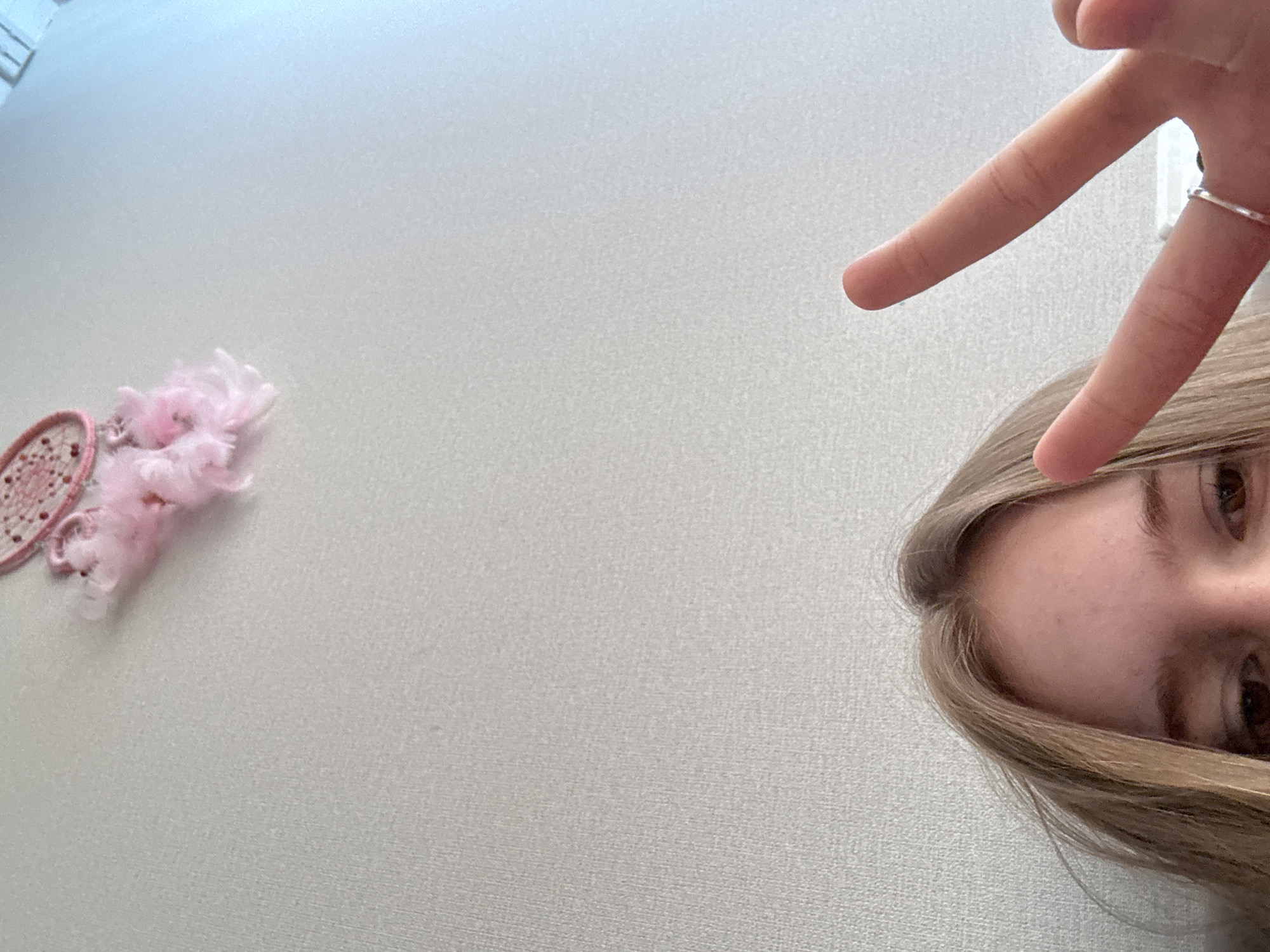 The image shows the upper portion of a young person's face and their hand making a peace sign. The person has blonde hair and brown eyes. They are wearing a silver ring on their right ring finger. In the background, there is a wall with a small pink dreamcatcher with feathers hanging from it. There is no indication of the location being Vsevolozhsk, Russia.