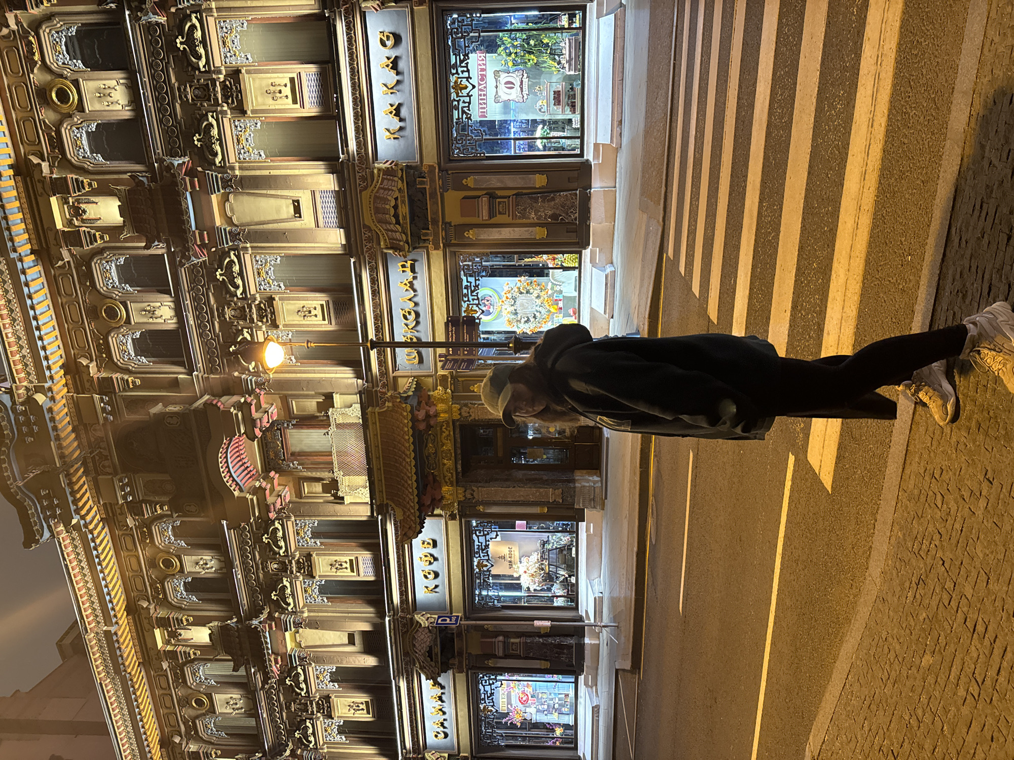 A medium-shot photograph taken at night shows a pedestrian, primarily from the back and side, crossing a zebra crossing. They are wearing dark clothing and light-colored sneakers. The location is identifiable as Myasnitskaya Street in Moscow, Russia, based on the distinctive architectural design of the buildings, characterized by ornate facades and detailed window displays. The storefronts feature signage in both Russian and English. The lighting is ambient, with streetlights illuminating the scene and reflections from the shops' window displays. The crosswalk stripes appear clear against the asphalt.