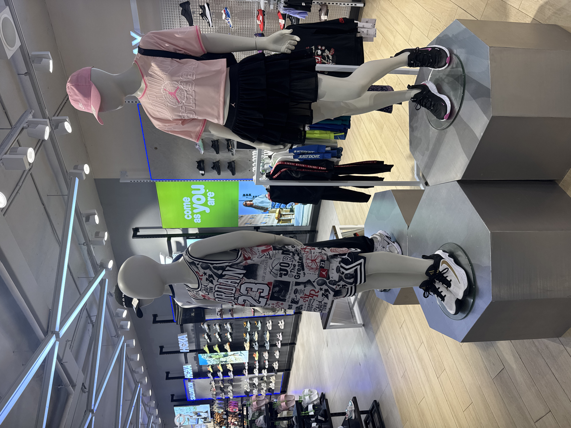 The image depicts the interior of a retail store, specifically located in Kuta, Indonesia. Two mannequins are prominently displayed on hexagonal pedestals. The mannequin on the left is wearing a Jordan brand basketball jersey and shorts, with white and black sneakers on its feet. The mannequin on the right is dressed in a pink cropped t-shirt and a black tiered skirt, also wearing black sneakers. Behind the mannequins, shelves are stocked with various athletic shoes and apparel. A large green sign with the text "COME AS YOU ARE" is visible on the back wall. The overall environment suggests a sportswear or athletic footwear store.