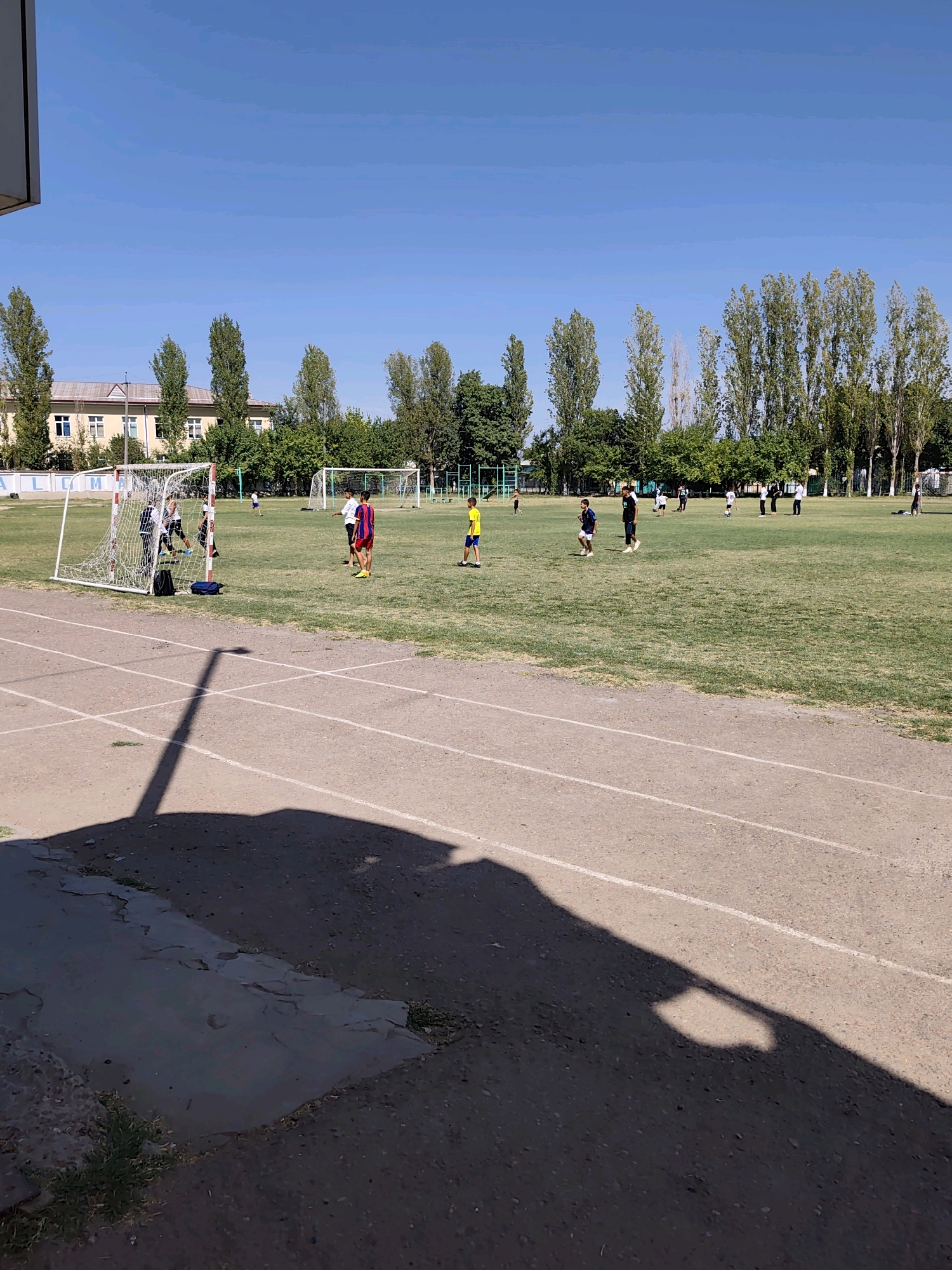 The image depicts a sunny day at a soccer field in the city of Malik, Uzbekistan. Several young boys are engaged in a soccer game on a well-maintained green field. A white soccer net is positioned on the left side of the frame, with some players gathered around it. Further into the field, other players are scattered, some with a ball, indicating an active match. In the background, a line of tall, leafy trees borders the field, with a building visible behind them. The foreground shows a portion of a running track with visible lane markings. The overall scene suggests a recreational activity taking place in a community park or school.