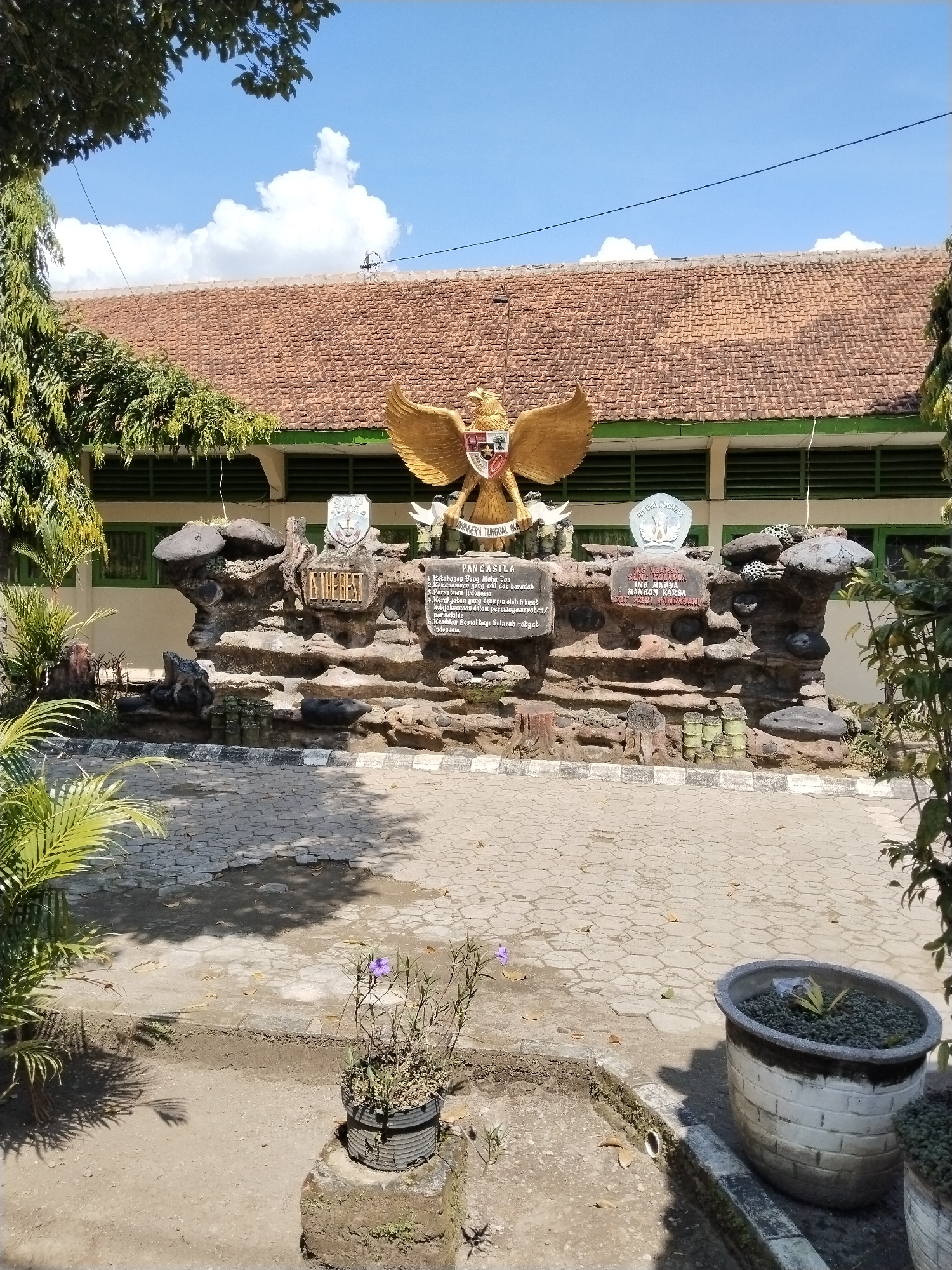 The image depicts a memorial or monument, likely in Sepat, Indonesia. It showcases a statue of Garuda Pancasila, the national emblem of Indonesia. Below the Garuda, a stone-like structure displays the Pancasila principles. The monument is positioned in front of a building with a brown tile roof and green window frames. In the foreground, there are potted plants on a paved area, partly shaded by trees. The sky is blue with scattered clouds.