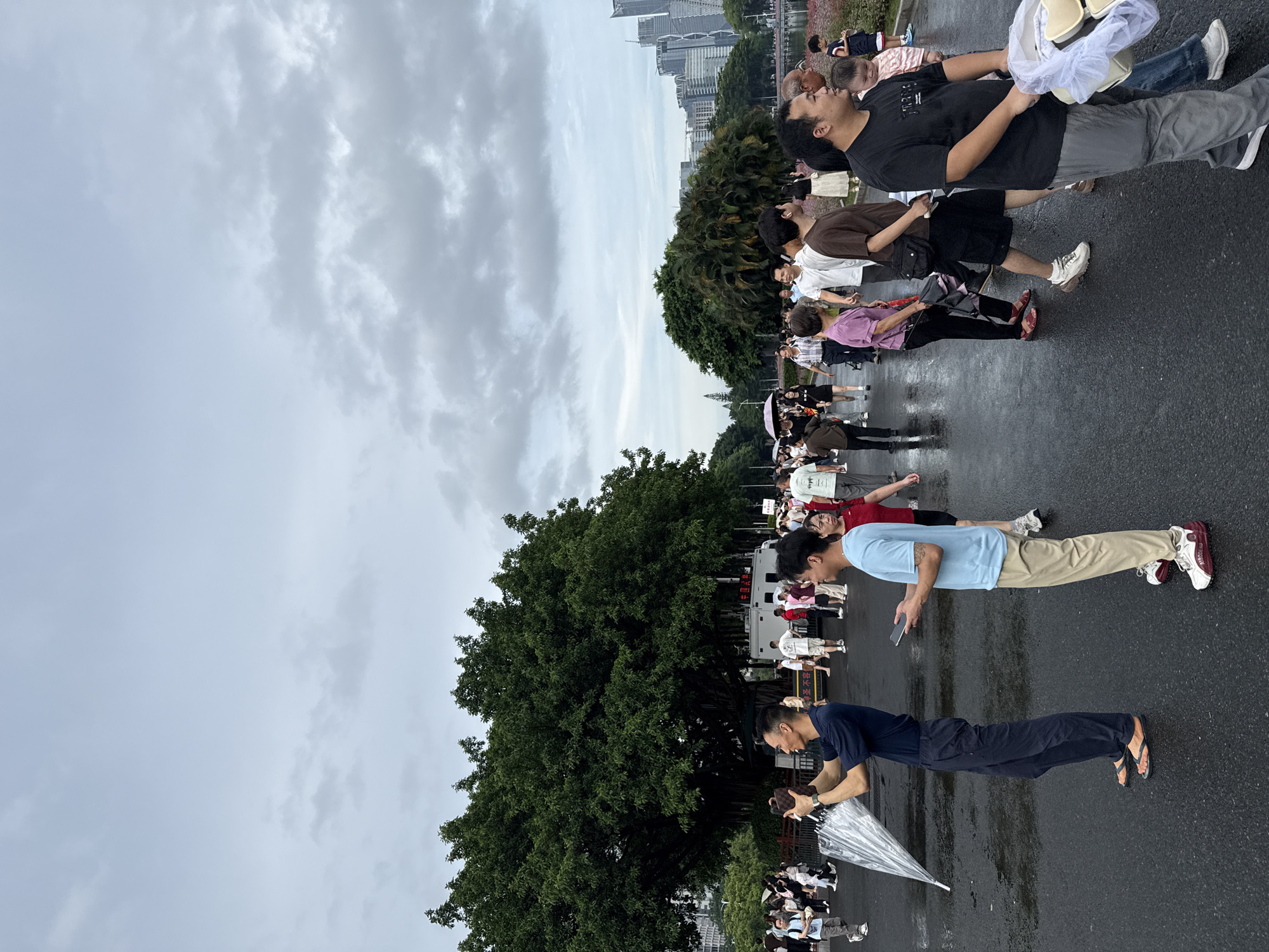 The image captures a street scene in Tianhe District, China, with a crowd of people walking on a wet asphalt surface. A large, leafy tree dominates the left side of the frame, with a white vehicle and more trees in the background. Buildings of various heights and architectural styles are visible in the distance, suggesting an urban environment. The sky is overcast with gray clouds.
In the foreground, several individuals are prominent. A man in a dark blue shirt and dark pants is holding a clear umbrella and a mobile device, appearing to take a photograph. To his right, another man in a light blue shirt and khaki pants holds a phone. Further to the right, a man in a black t-shirt and gray pants is walking, carrying a white plastic bag containing some items. Other people, including men, women, and children, are dispersed throughout the scene, some walking individually, others in groups. Some individuals are carrying umbrellas, indicating recent or ongoing rain. The wet pavement reflects the surrounding environment.