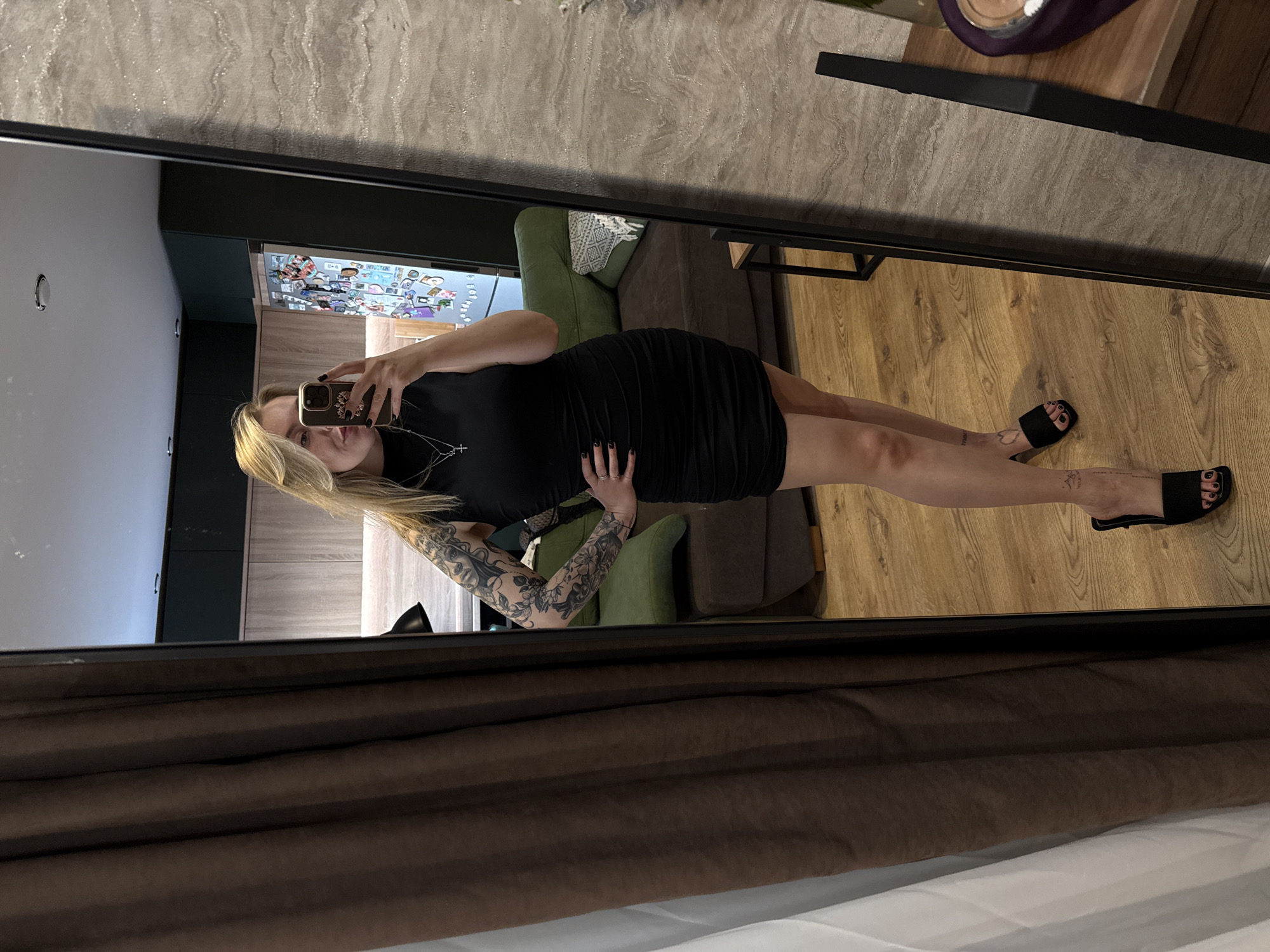 A woman is reflected in a full-length mirror indoors. She has blonde hair and is wearing a black dress and black sandals. Tattoos are visible on her arms. She is holding a smartphone, presumably taking the photo. The background includes a sofa, a wooden floor, and a glimpse of a kitchen area with a refrigerator covered in magnets. The location appears to be an apartment or home. There's no readily discernible information to confirm the location in Krasnoyarsk, Russia.