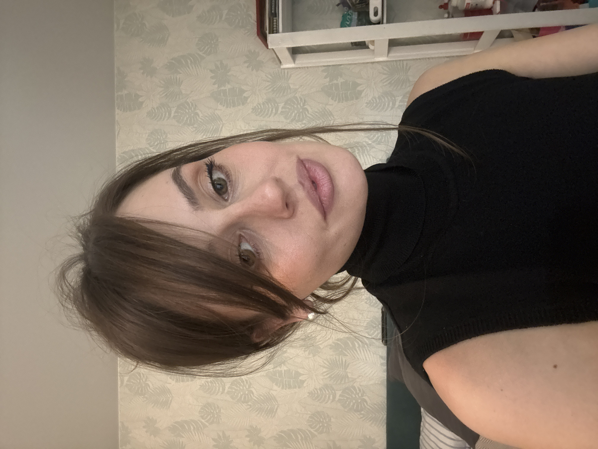 An adult female is depicted in a medium-shot portrait, angled slightly to her left, looking towards the camera. She has brown hair styled with a fringe and some loose strands, and light-toned skin. Her eyes are greenish-brown, with visible eye makeup including eyeliner and mascara, and her lips are painted with a light pink gloss. A small white stud earring is present in her visible right ear. She is wearing a black sleeveless turtleneck top. The background indicates an indoor domestic setting. A wall behind her features light-colored wallpaper with a repeating pattern of stylized leaf motifs. To the left, a portion of a bed or sofa with a dark green headboard or cushion and striped white and grey bedding is visible. To her right, a white multi-shelf storage unit or bookcase contains various items, including a red object on the top shelf and bottles on lower shelves.
