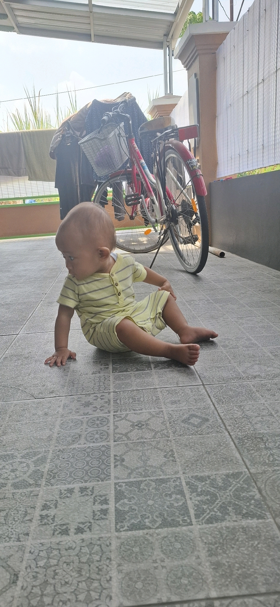 In Desa Gagaksipat, Indonesia, a bald baby wearing a light yellow and grey striped onesie sits on a tiled floor. The baby is positioned facing the left side of the frame, with their right arm resting on their right leg and their left hand touching the floor. Their legs are bent, and their feet are visible. In the background, a red bicycle with a basket is propped up, leaning against a structure. The environment appears to be outdoors, under a covered area, with sunlight casting shadows.